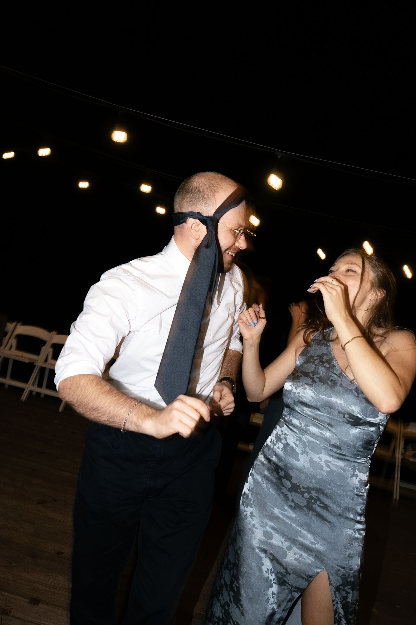 A bald man playfully covers his face with a tie while dancing with a laughing woman in a silver dress at an evening event.