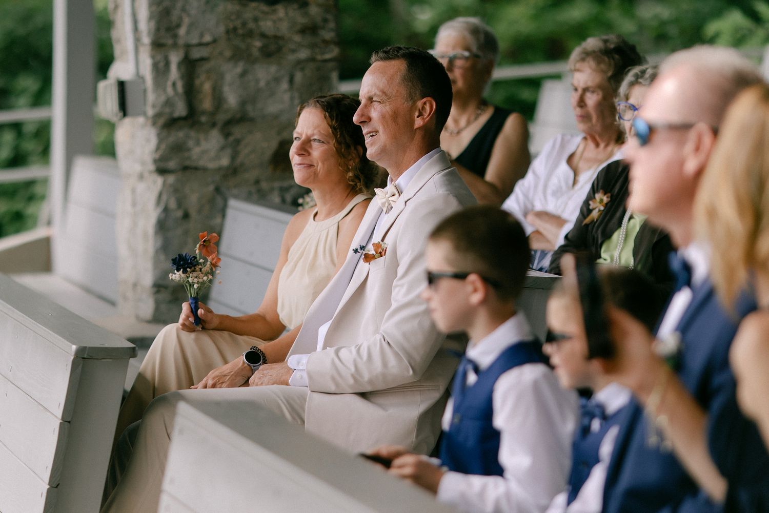 Wedding guests seated outdoors, smiling couple in foreground, woman holding bouquet, man in tan suit, stone backdrop.