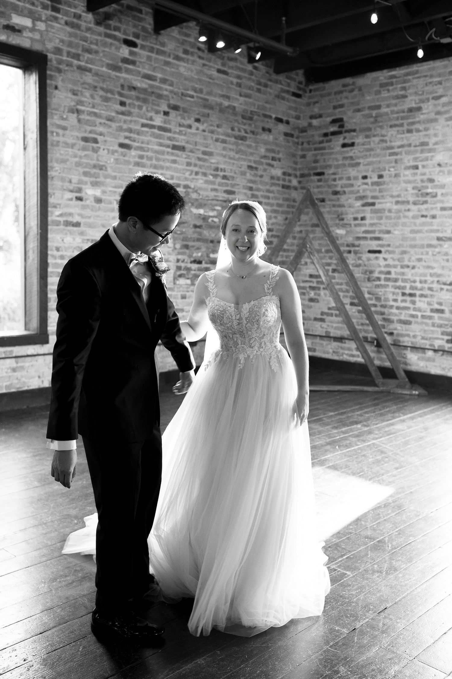 A bride and groom share an intimate moment in a rustic brick-walled venue with natural light streaming through windows.