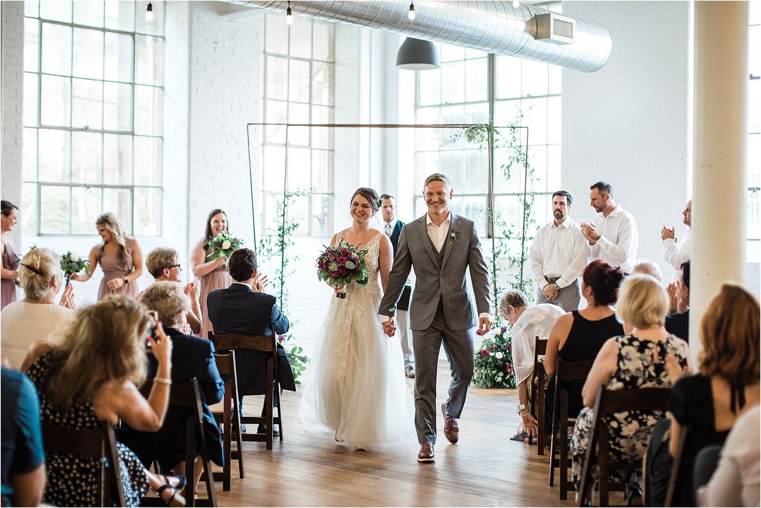 A couple walks down the aisle while wedding guests capture photos in a bright, modern venue.