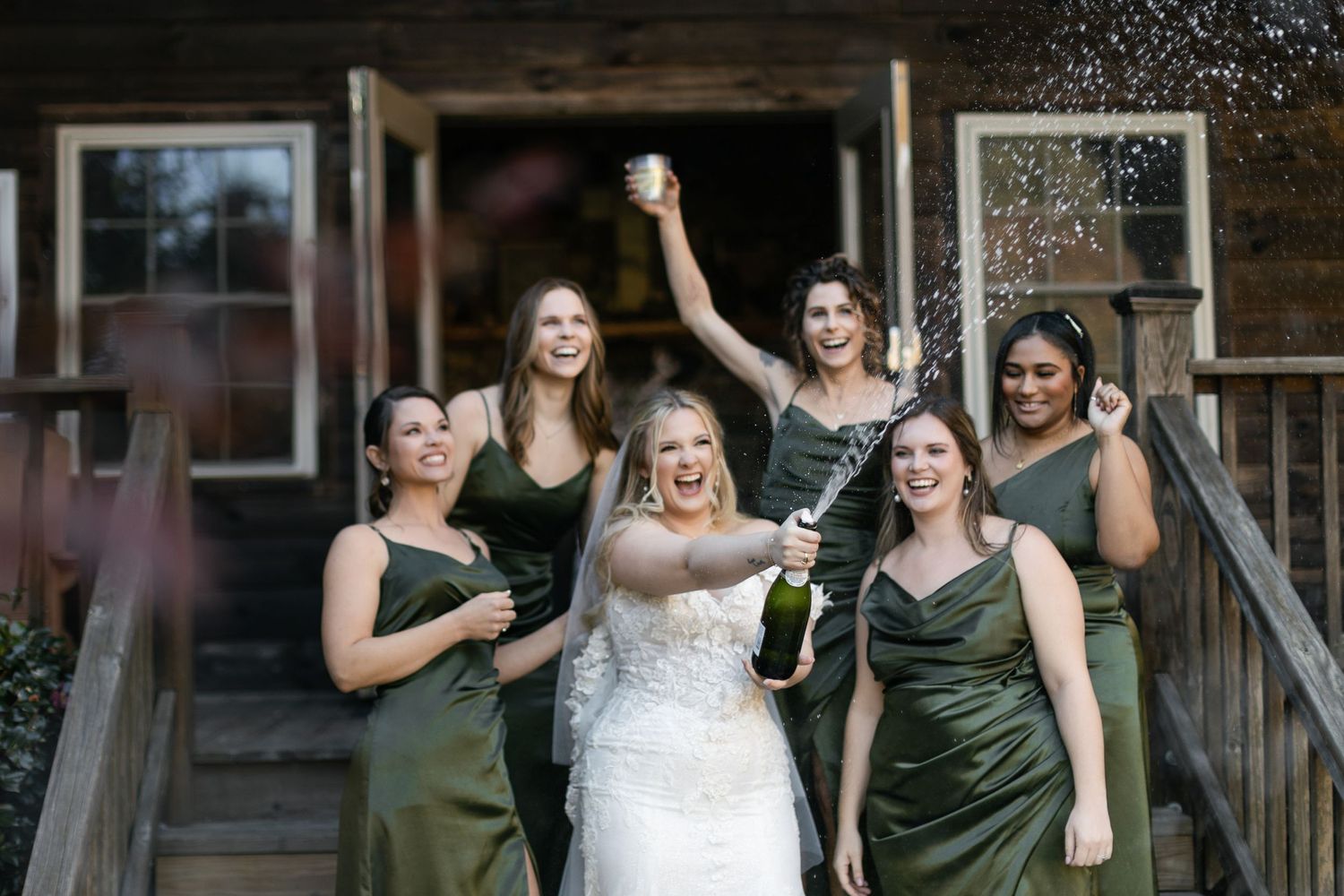 A group in matching green dresses celebrates with a champagne spray outside a rustic wooden building.