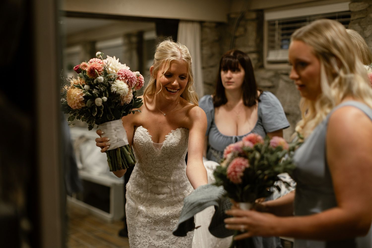 A bride in a lace strapless gown holds a pink and white bouquet while celebrating with her bridesmaids indoors.