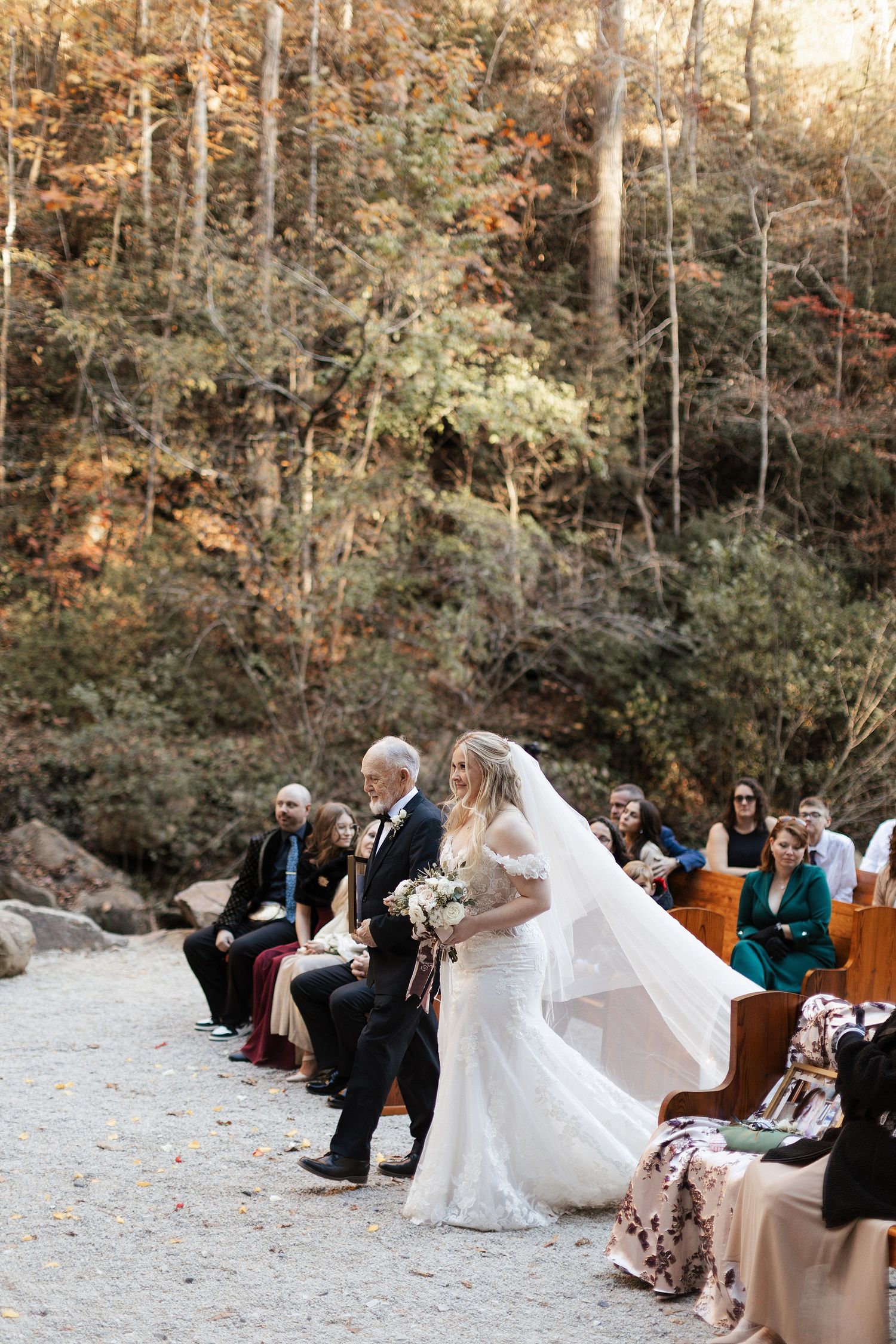 An outdoor autumn wedding ceremony setup with wooden chairs and colorful fall foliage in the forest background.