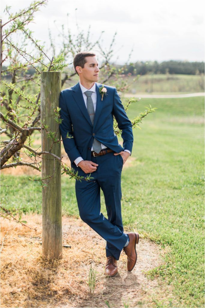 Groom in navy suit and brown shoes poses casually against rustic fence post.