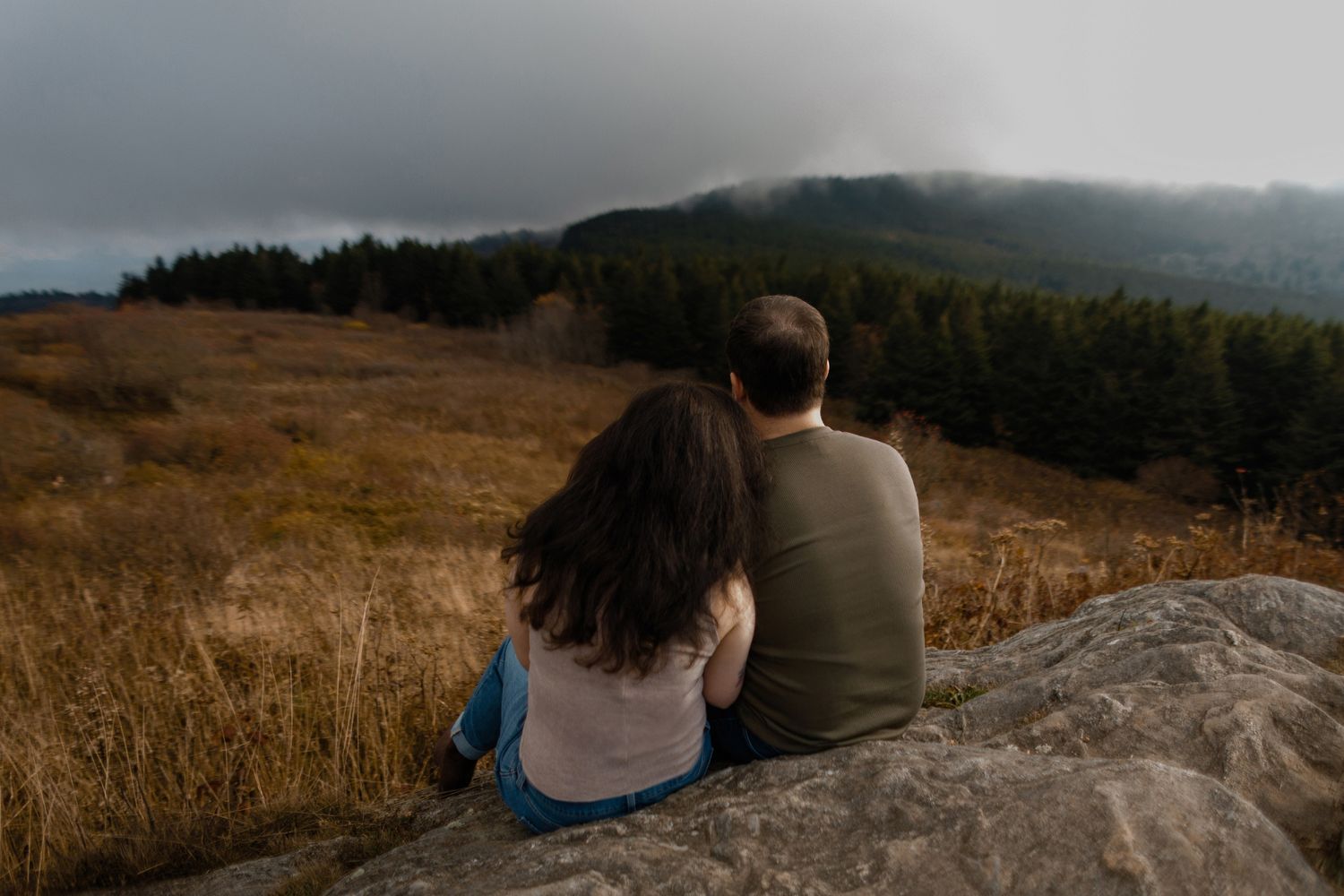 Couple sits together on large rock overlooking golden hillside at sunset with mountains in background.