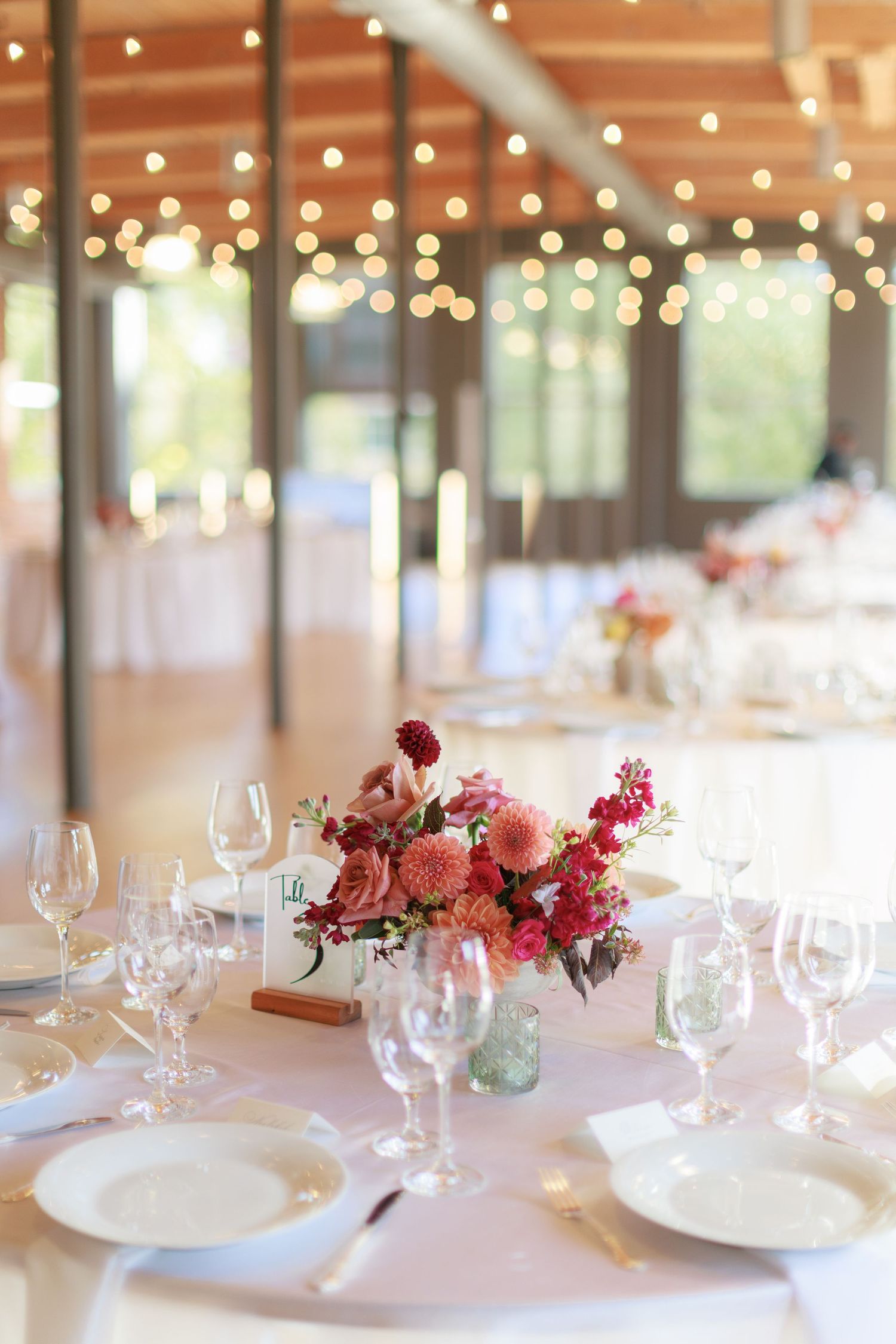 Elegant wedding reception table with pink floral centerpiece, crystal glassware, white plates, and soft string lights in background.
