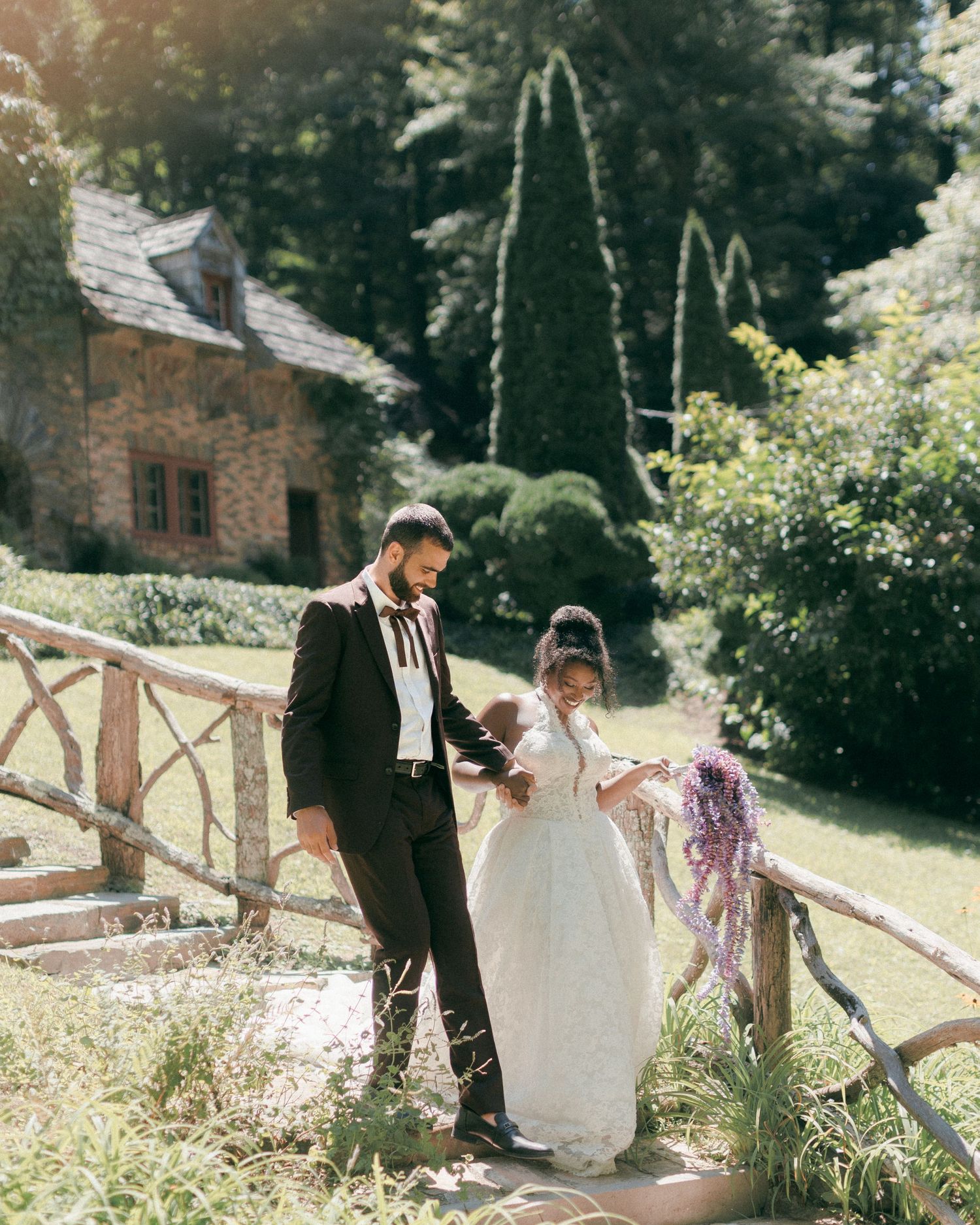 A bride in a white gown and groom in a black suit walk hand in hand on a rustic wooden bridge in a lush garden.