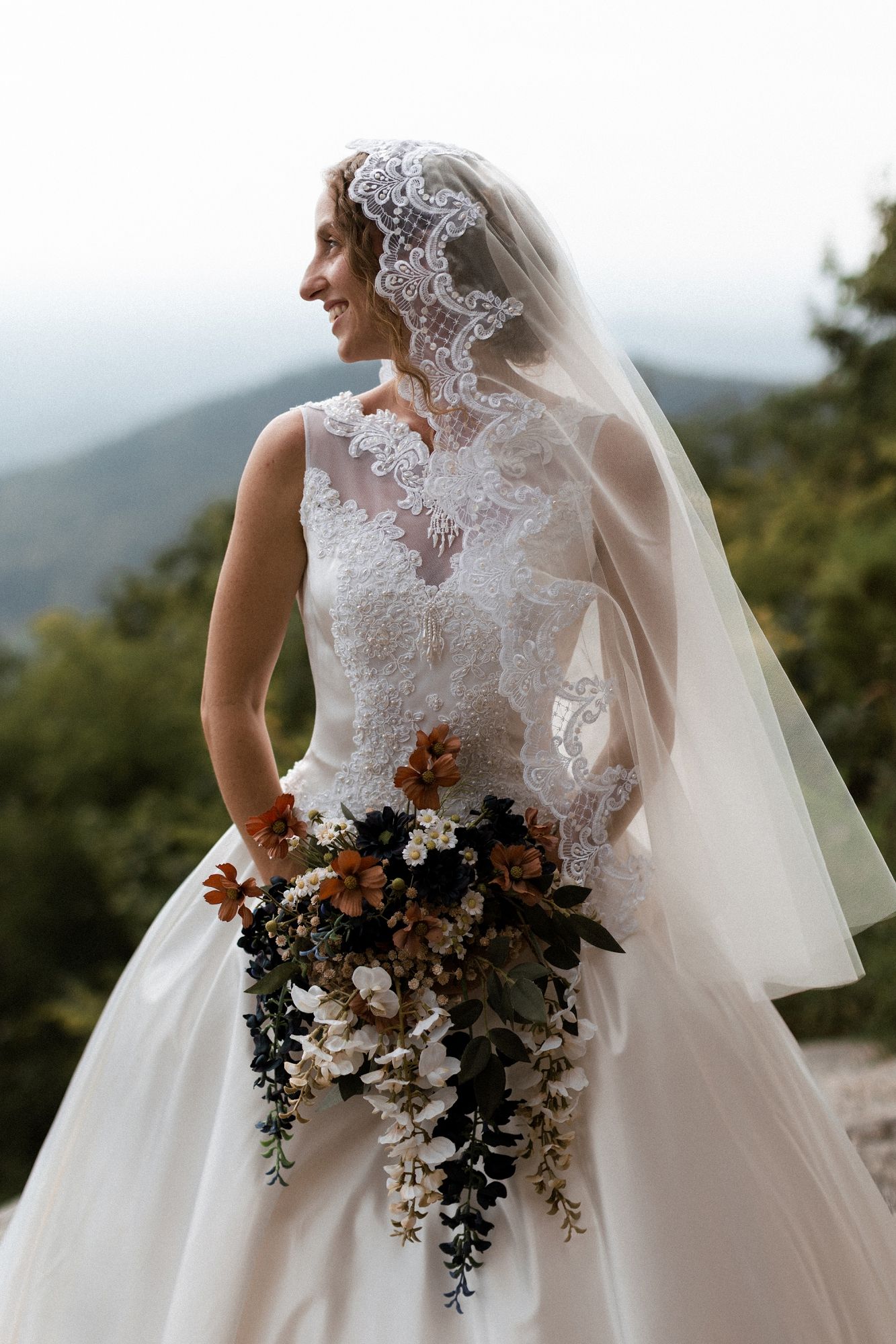 Bride in lace wedding gown and veil holds autumn bouquet with mountain backdrop.