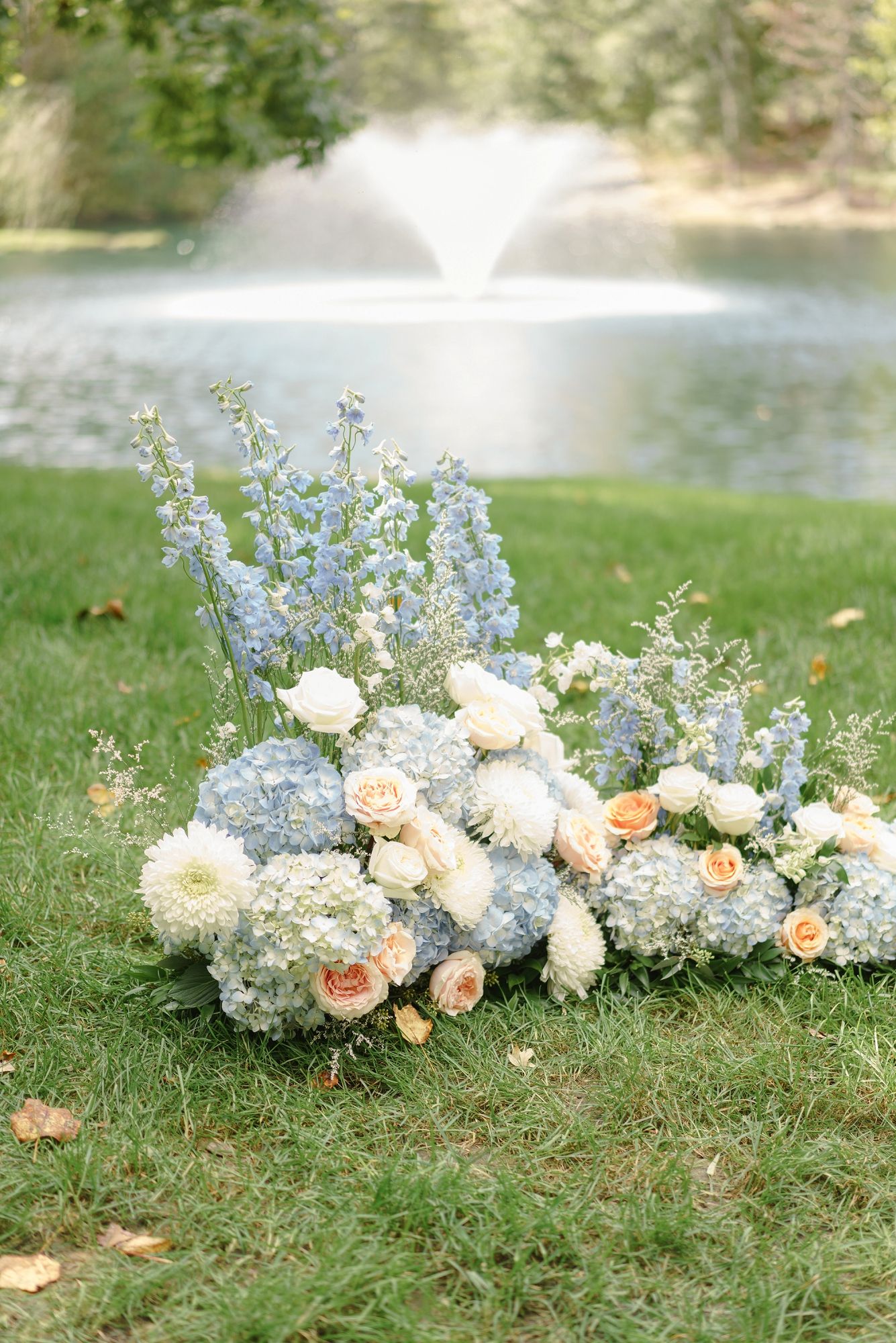 Elegant wedding floral arrangement of blue hydrangeas, peach roses, and delphiniums on grass near fountain.