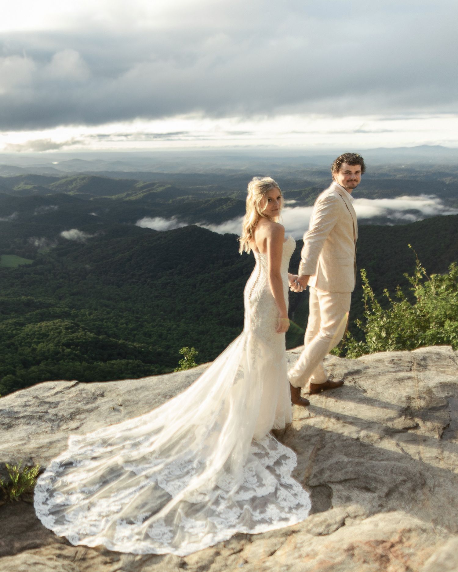 A couple in wedding attire stands on a mountaintop overlooking a misty valley with dramatic cloudy skies at sunset.