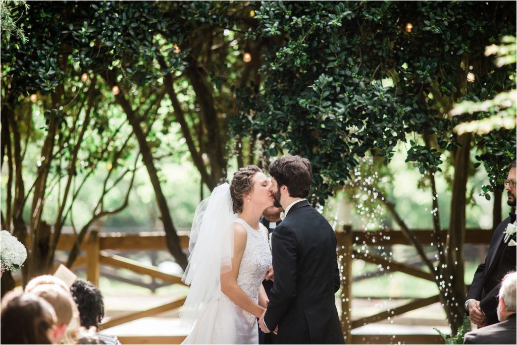 Romantic outdoor wedding kiss moment beneath a canopy of sunlit trees.