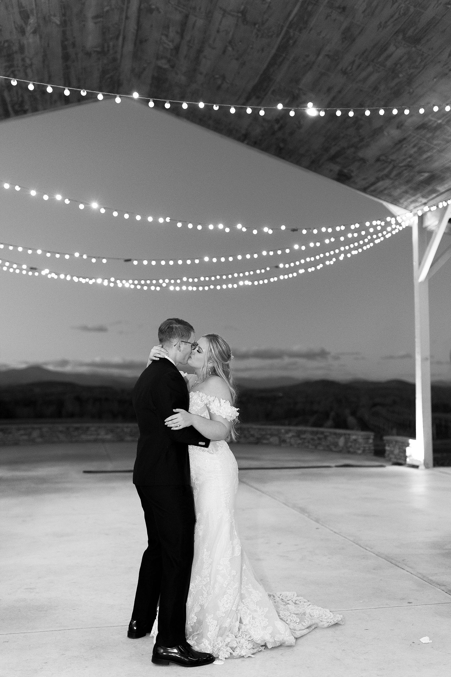 A romantic couple shares their first dance under twinkly string lights at an outdoor wedding reception venue at dusk.