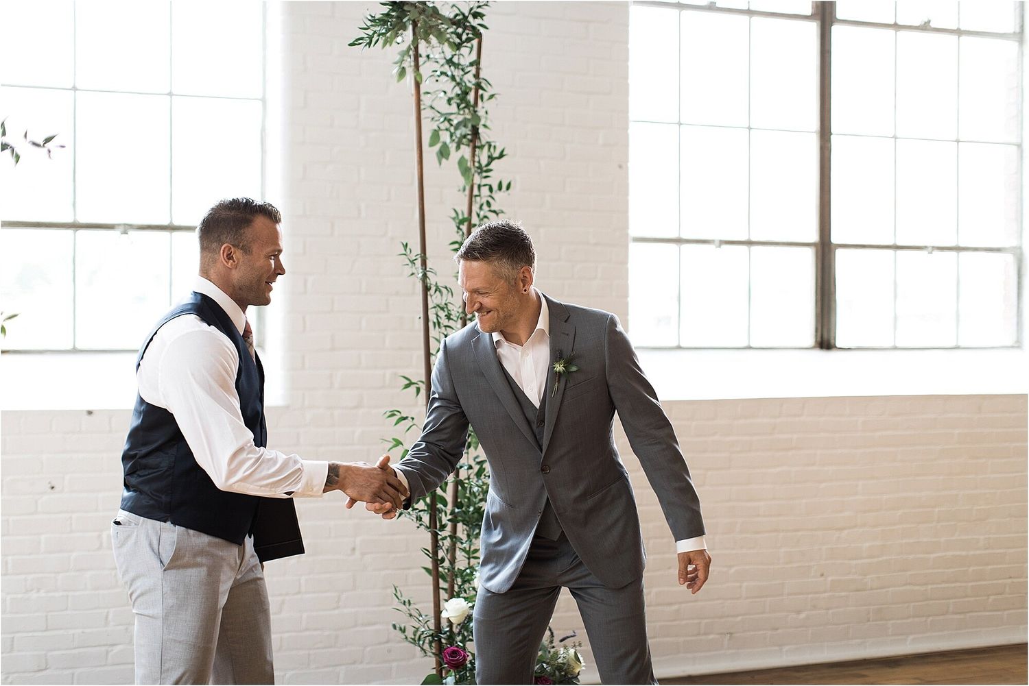 Two people in formal attire shake hands in a bright indoor space with white walls.