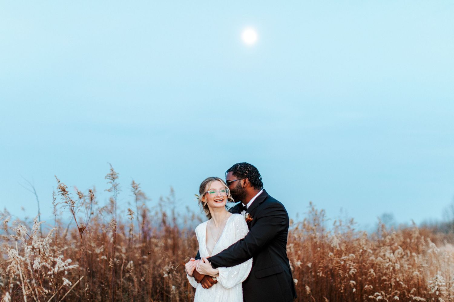 A couple shares a tender moment in a field of tall grass under a pale blue sky.