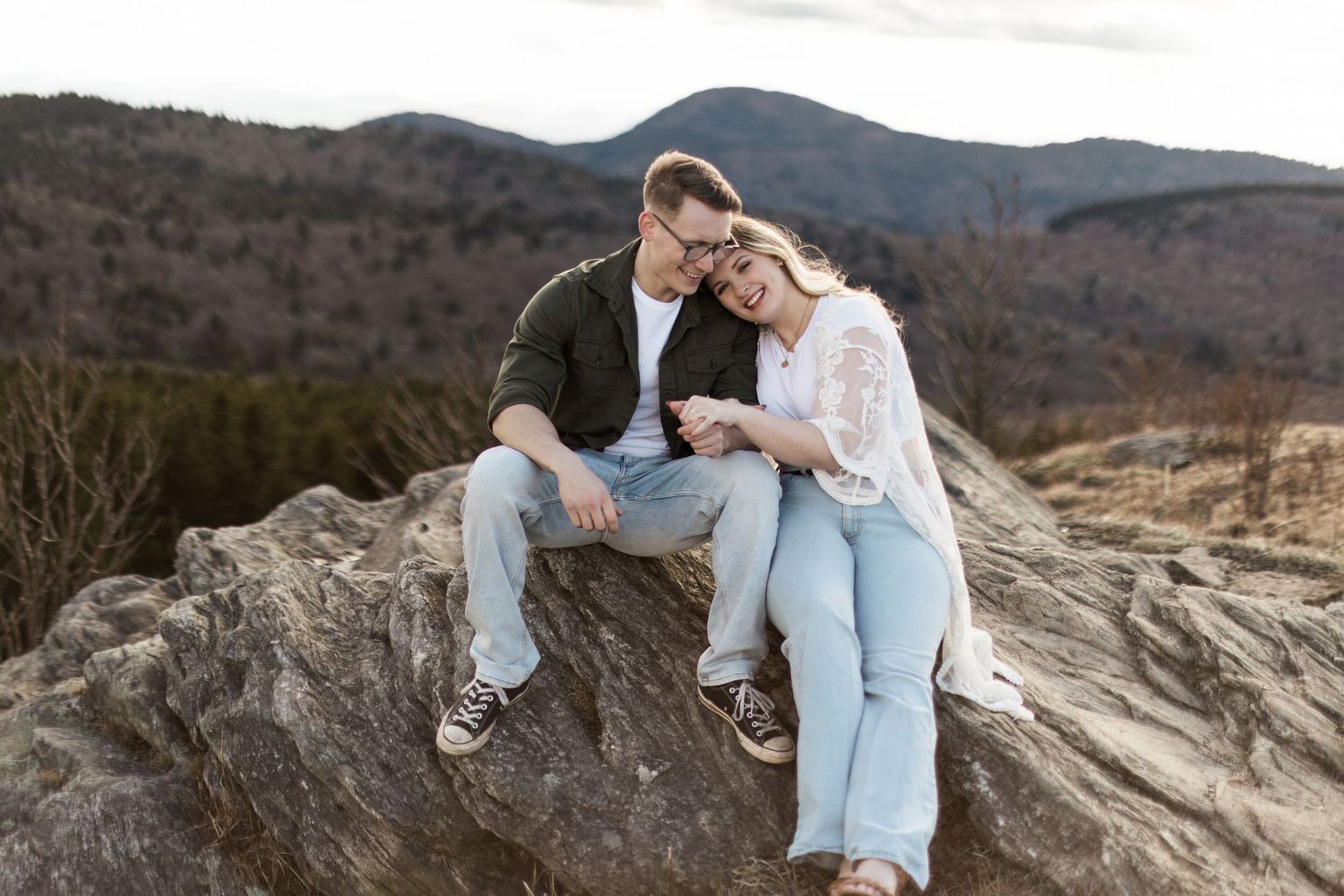 A couple sits close together on a large rock formation overlooking mountains during sunset.