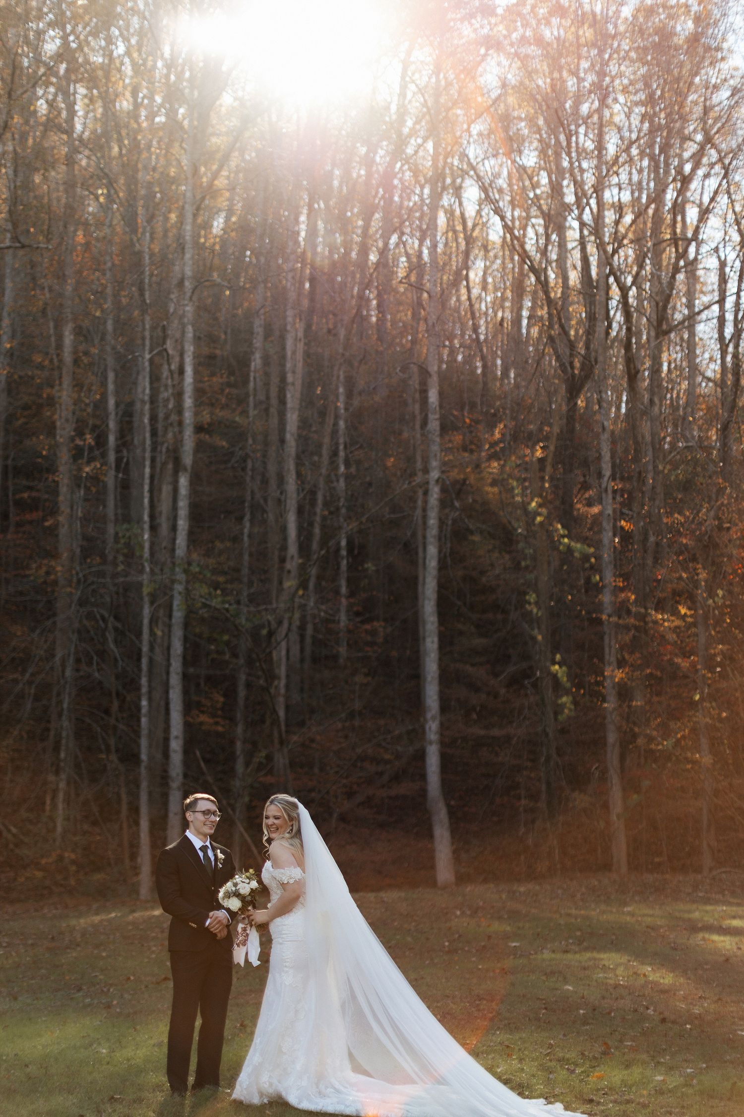 A couple in wedding attire stands together in a sunlit autumn forest with tall bare trees and golden foliage.