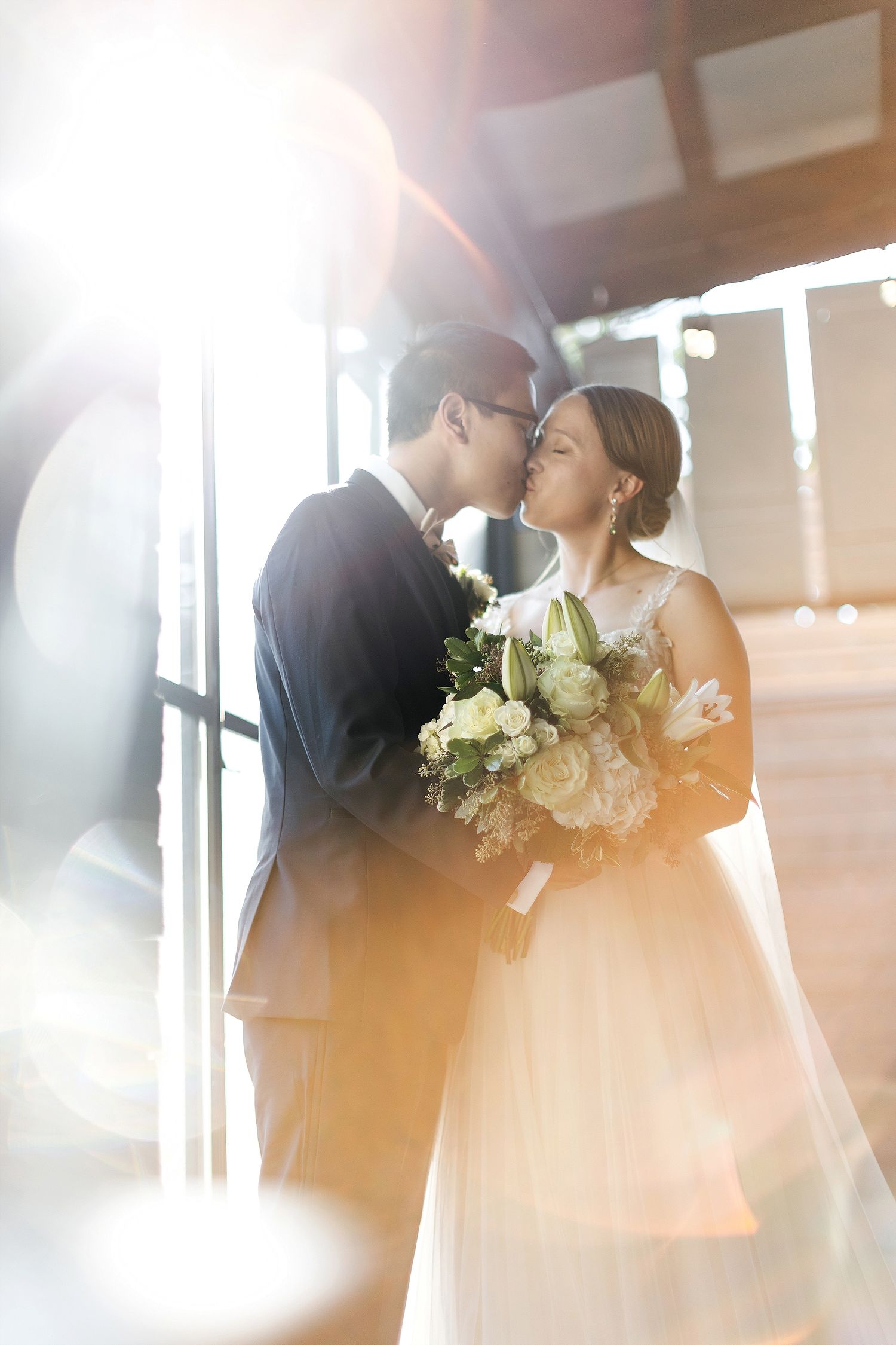 Newlywed couple in romantic bridal portrait sharing an intimate moment in sunlit window setting with white bouquet.