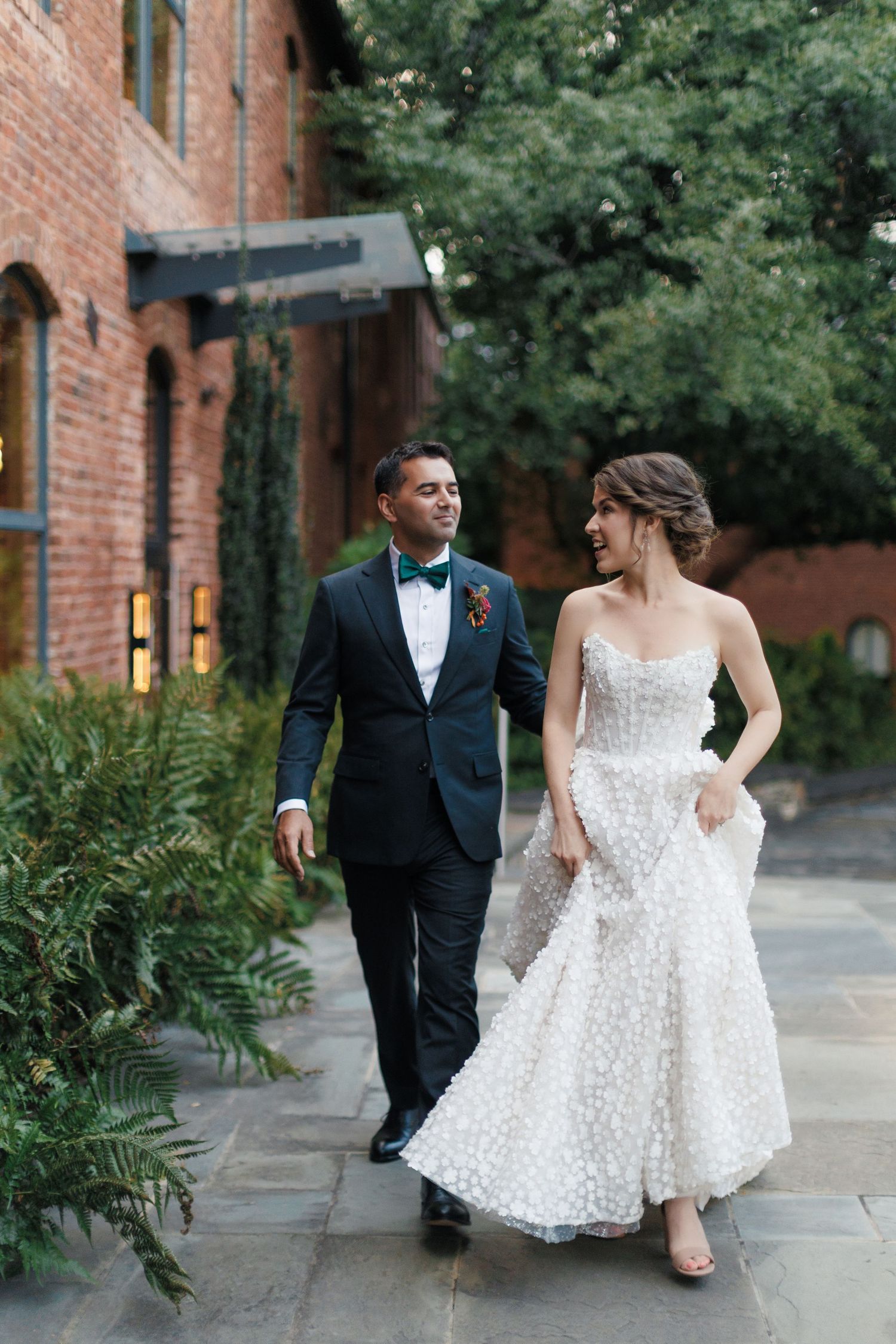 A bride in a white floral gown and groom in a dark tuxedo walk together smiling outside the Huguenot Mill and Loft in Greenville, SC.