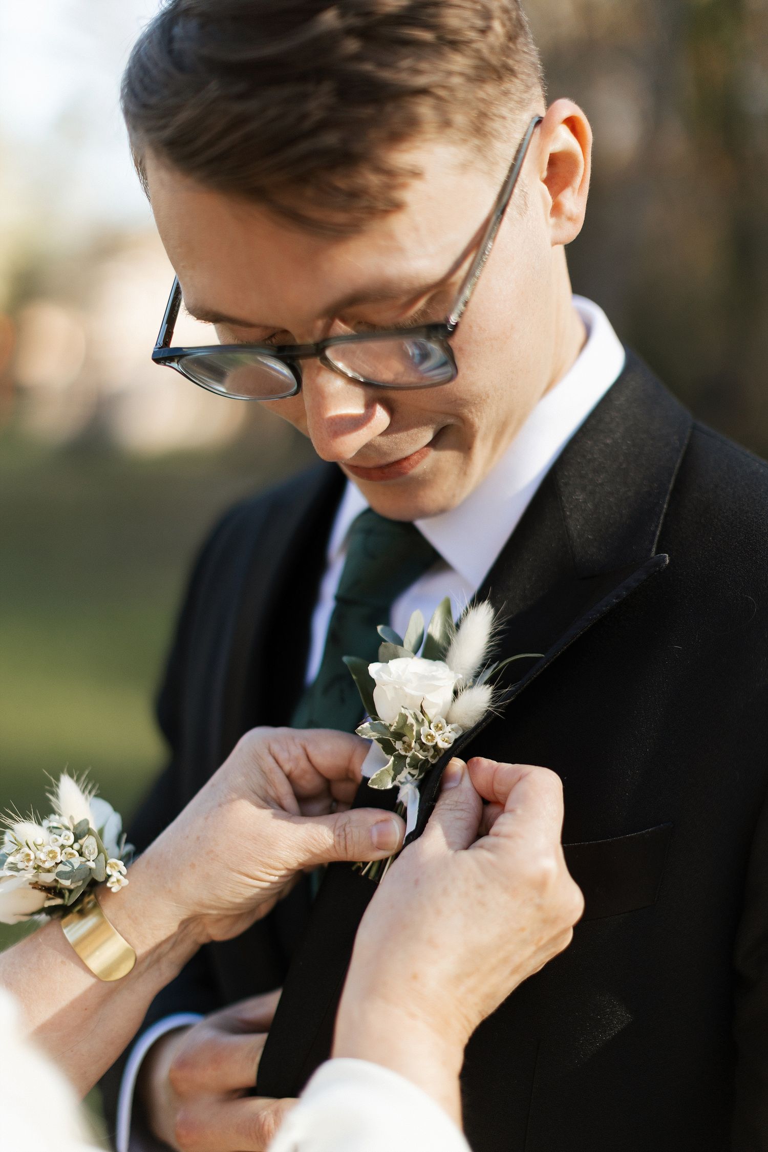Someone helps pin a white boutonniere flower to the lapel of a groom wearing glasses and a black suit at an outdoor wedding.