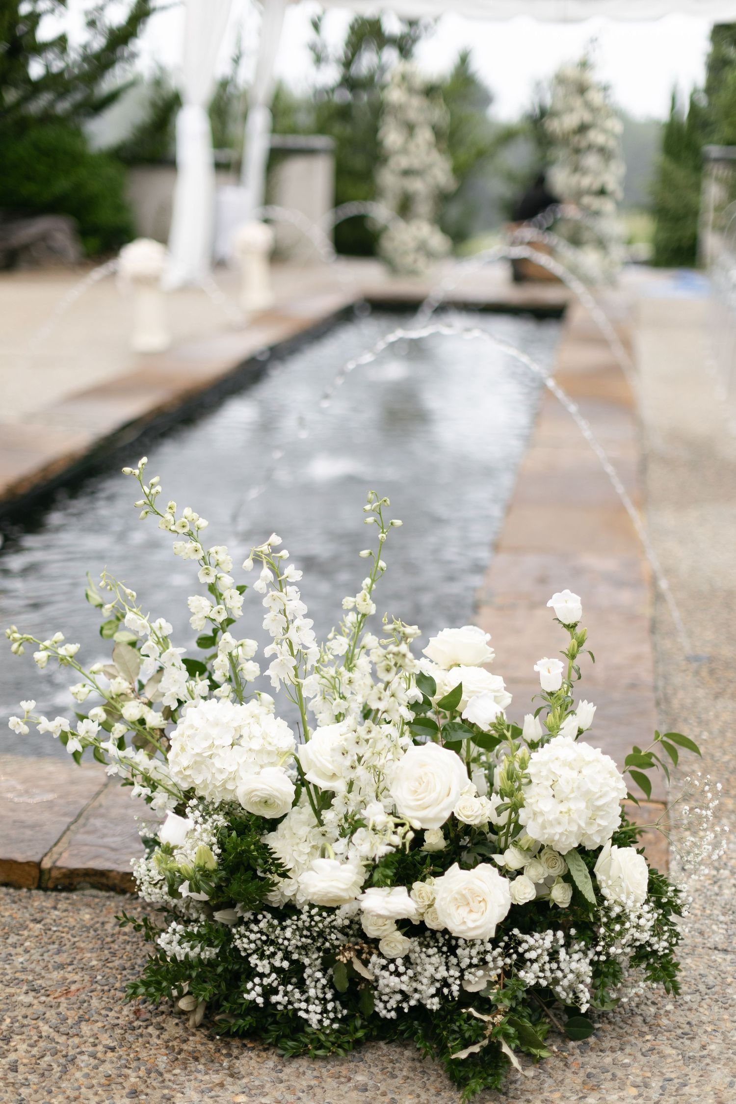 White floral arrangement with roses and delicate blooms sits by an elegant reflective pool with water features.