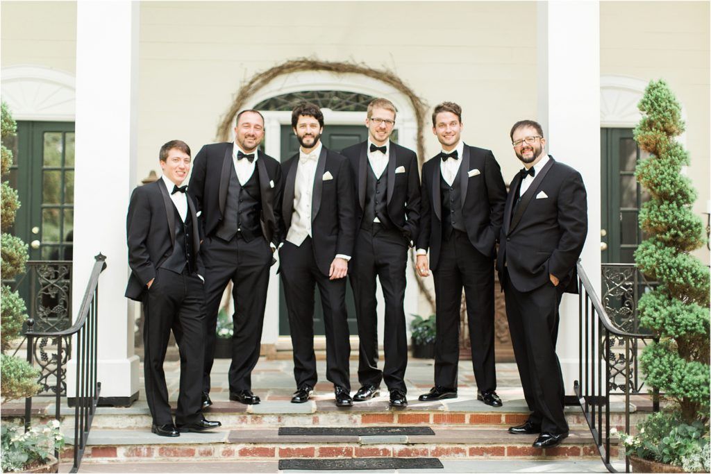 Groomsmen in black tuxedos pose on white columned entrance steps.