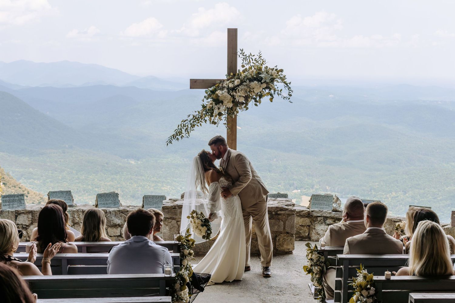 A scenic mountain wedding ceremony takes place on an outdoor terrace with guests seated and a decorated cross in the background.