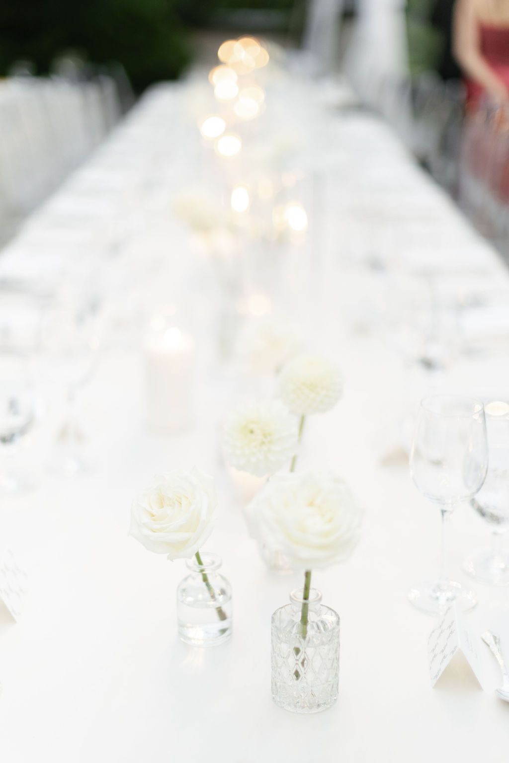 White roses in small glass vases create an elegant table setting with twinkling lights in the background.