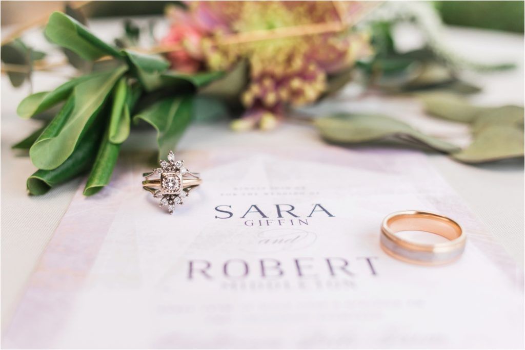 Wedding rings and invitation with names Sara and Robert displayed with greenery and flowers.