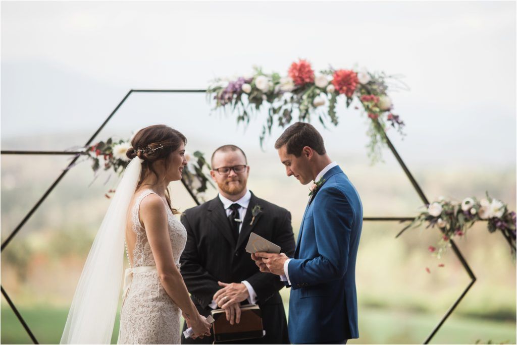 Outdoor wedding ceremony under a hexagonal arch decorated with pink and white flowers against a mountain backdrop.