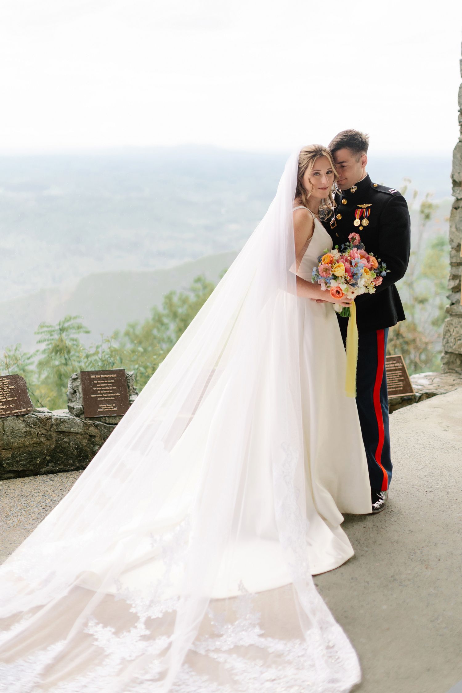 A bride in a flowing white gown and groom in military dress uniform embrace while posing on a scenic overlook.