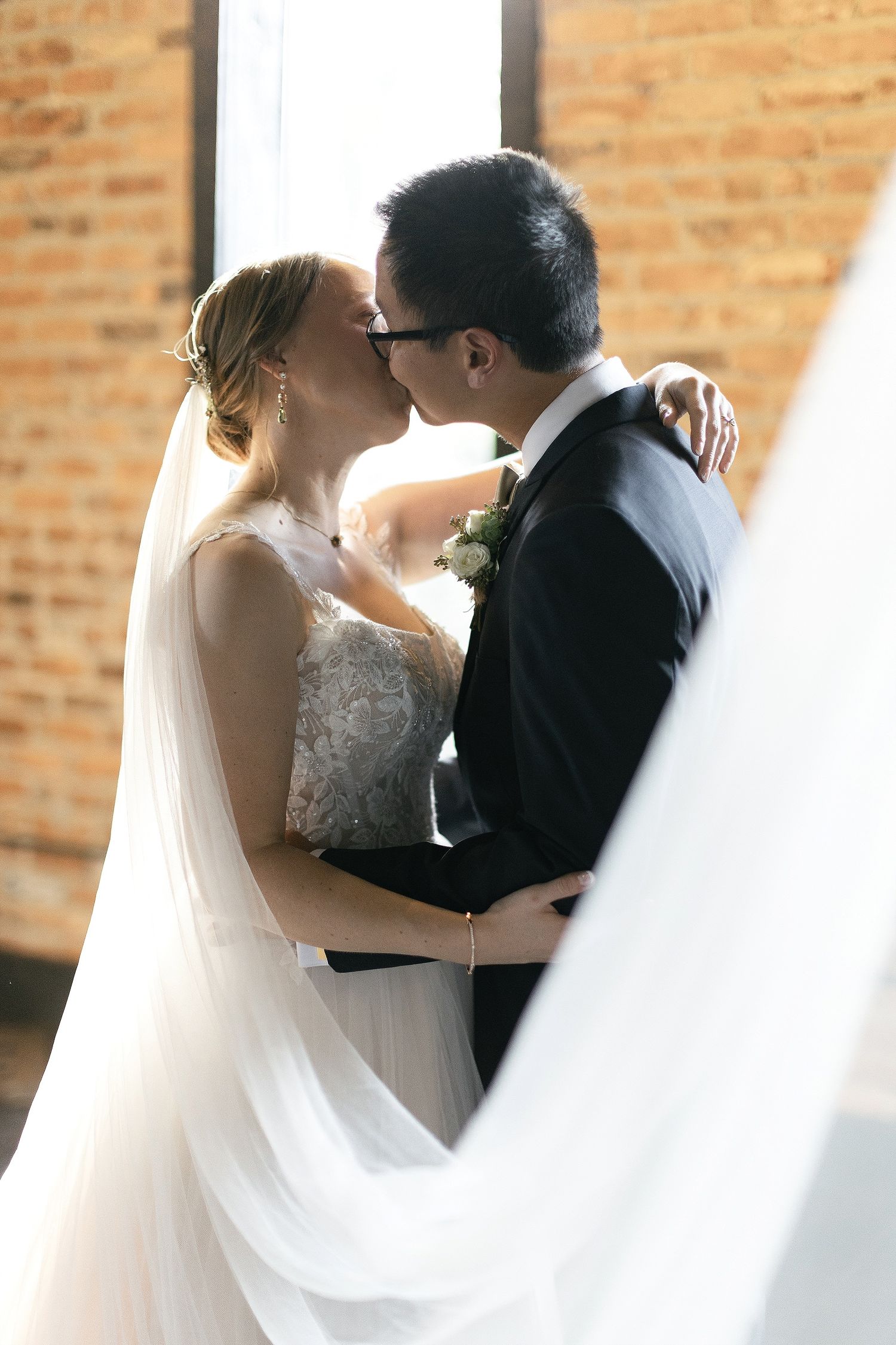 A romantic wedding moment with a bride in lace dress and flowing veil embracing her new spouse against a brick wall backdrop.