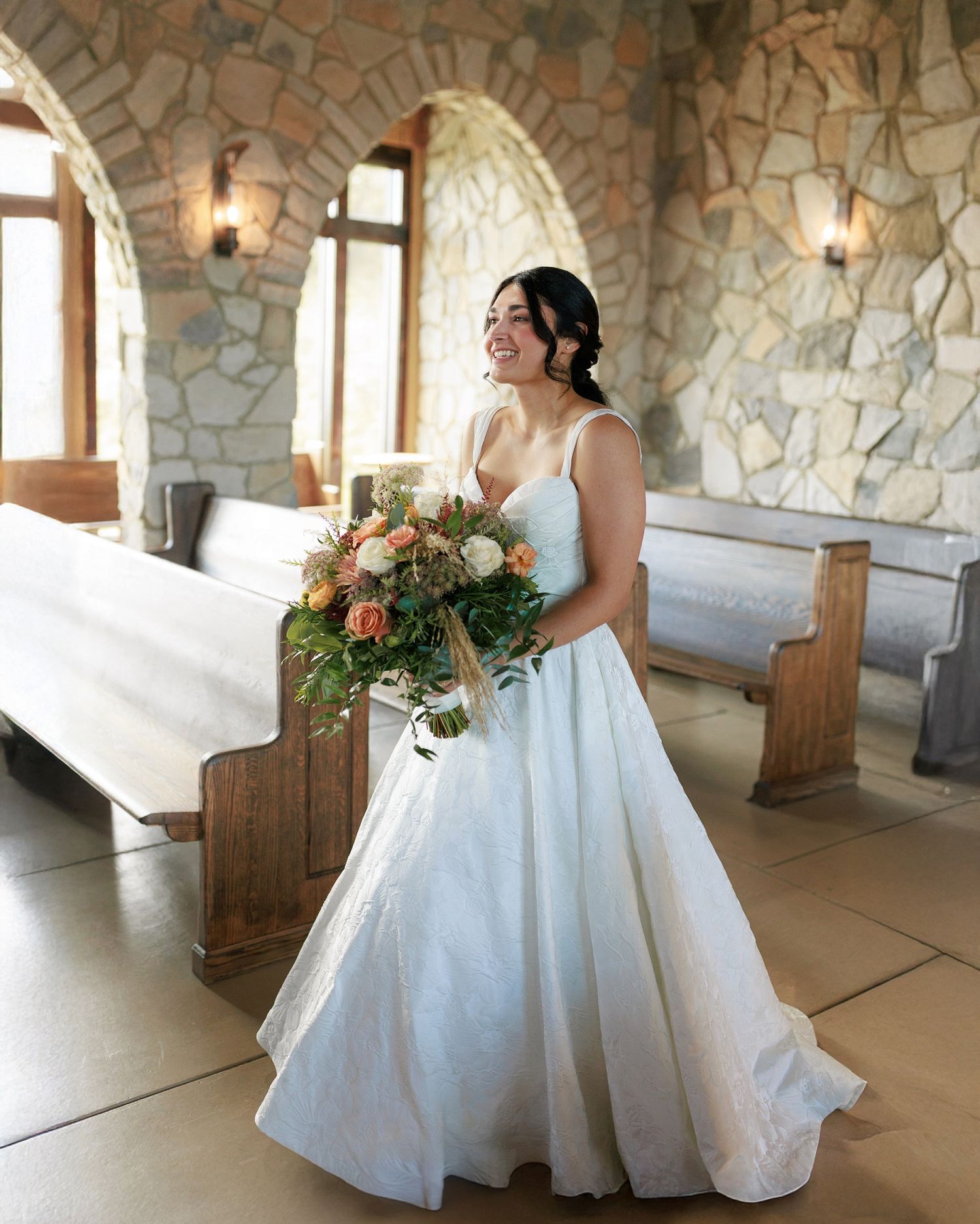 Bride in elegant white ballgown twirls playfully in a stone-walled venue with arched windows and natural lighting.
