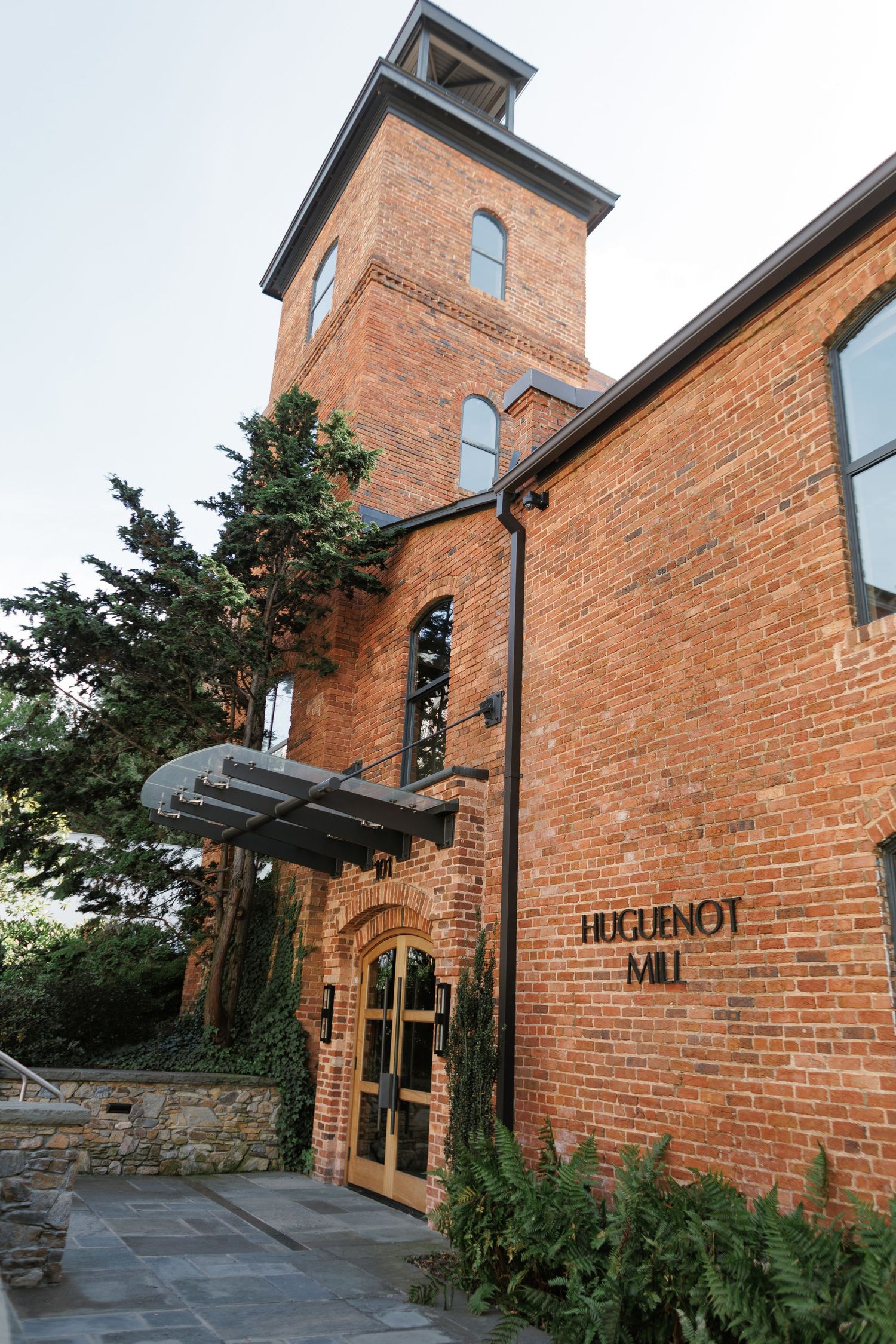 Historic Huguenot Mill red brick building with bell tower, wooden entrance doors, and lush greenery in a scenic outdoor setting.