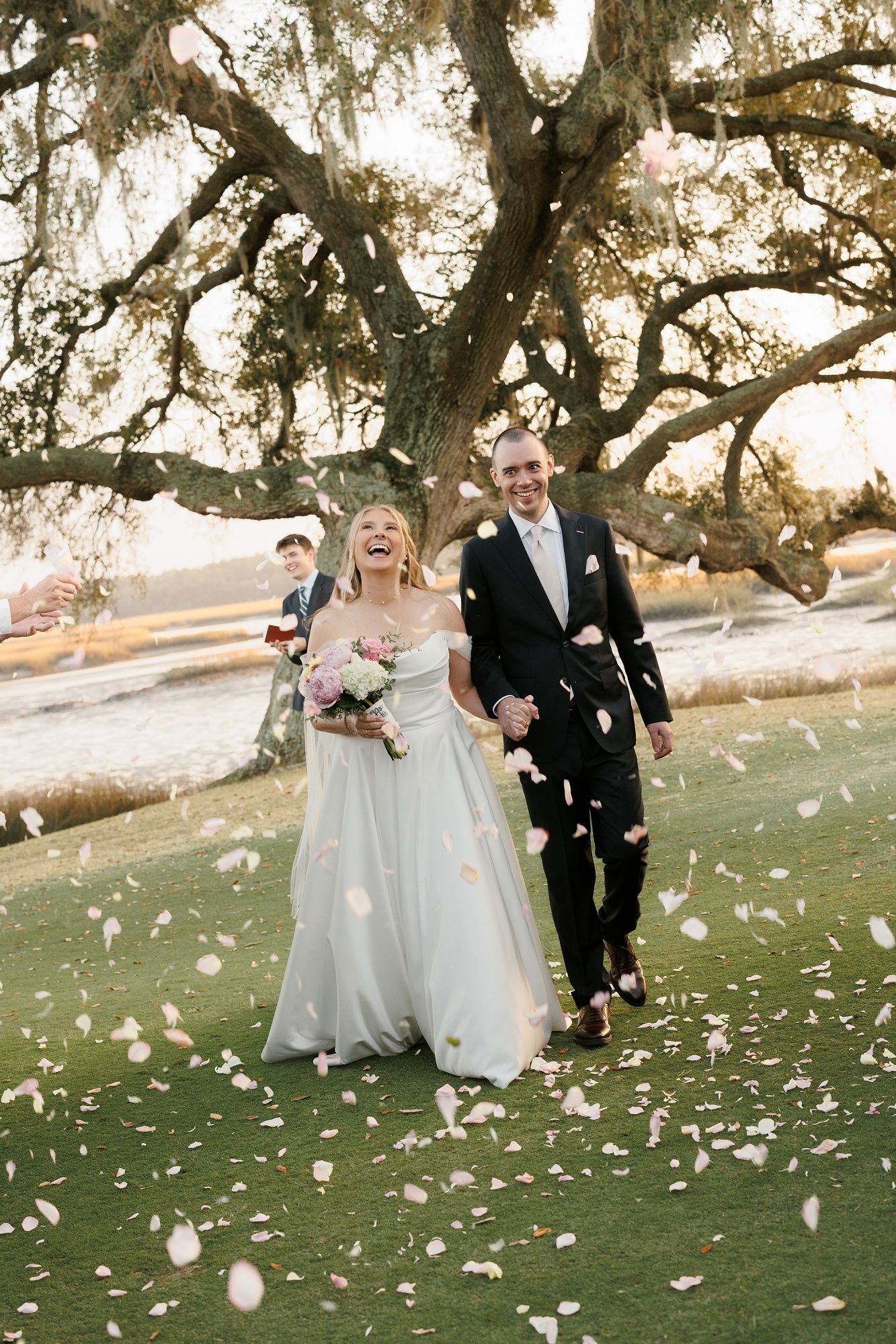 A joyful wedding couple walks on grass under a majestic oak tree while flower petals fall around them.
