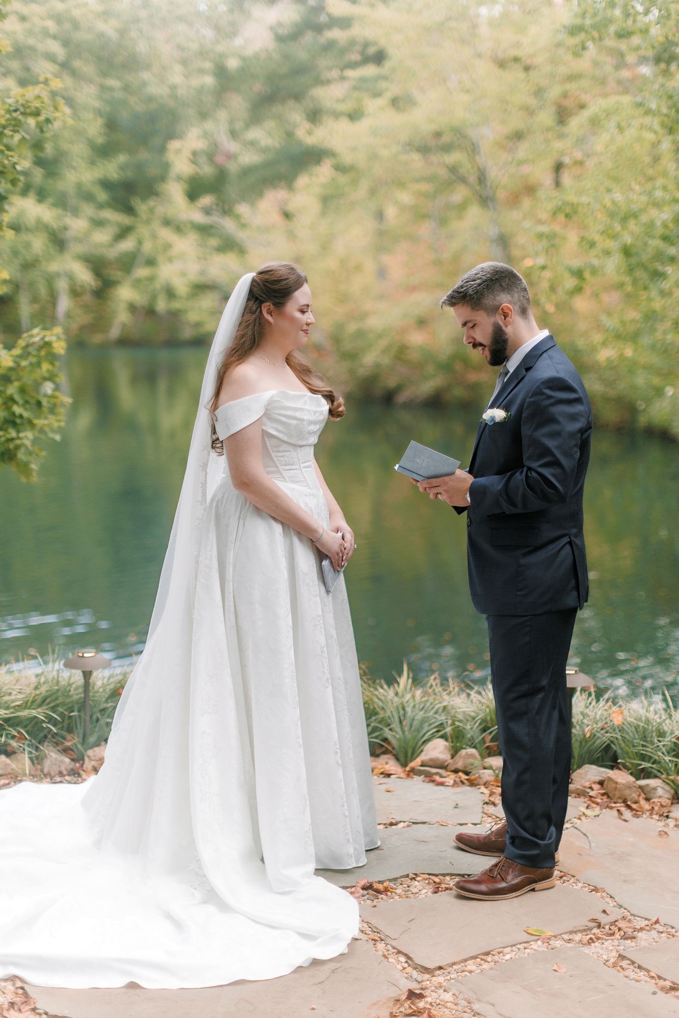 Bride in white off-shoulder gown and groom in navy suit exchange vows outdoors by a serene pond.