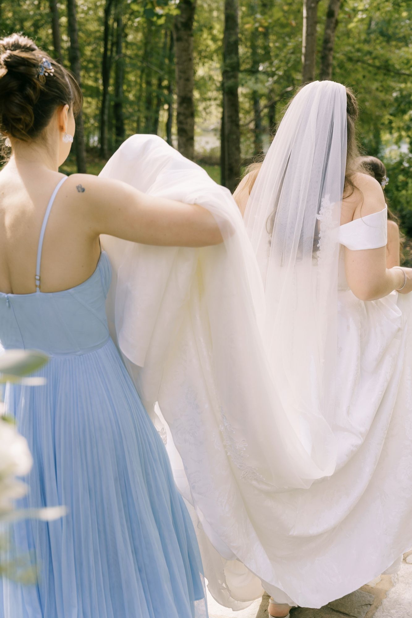 Bridesmaid in blue dress helps carry bride's white wedding gown train outdoors in lush green forest setting.