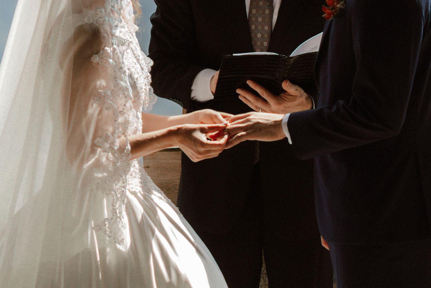 Groom placing wedding ring on bride's finger during outdoor ceremony, bride in lace dress.