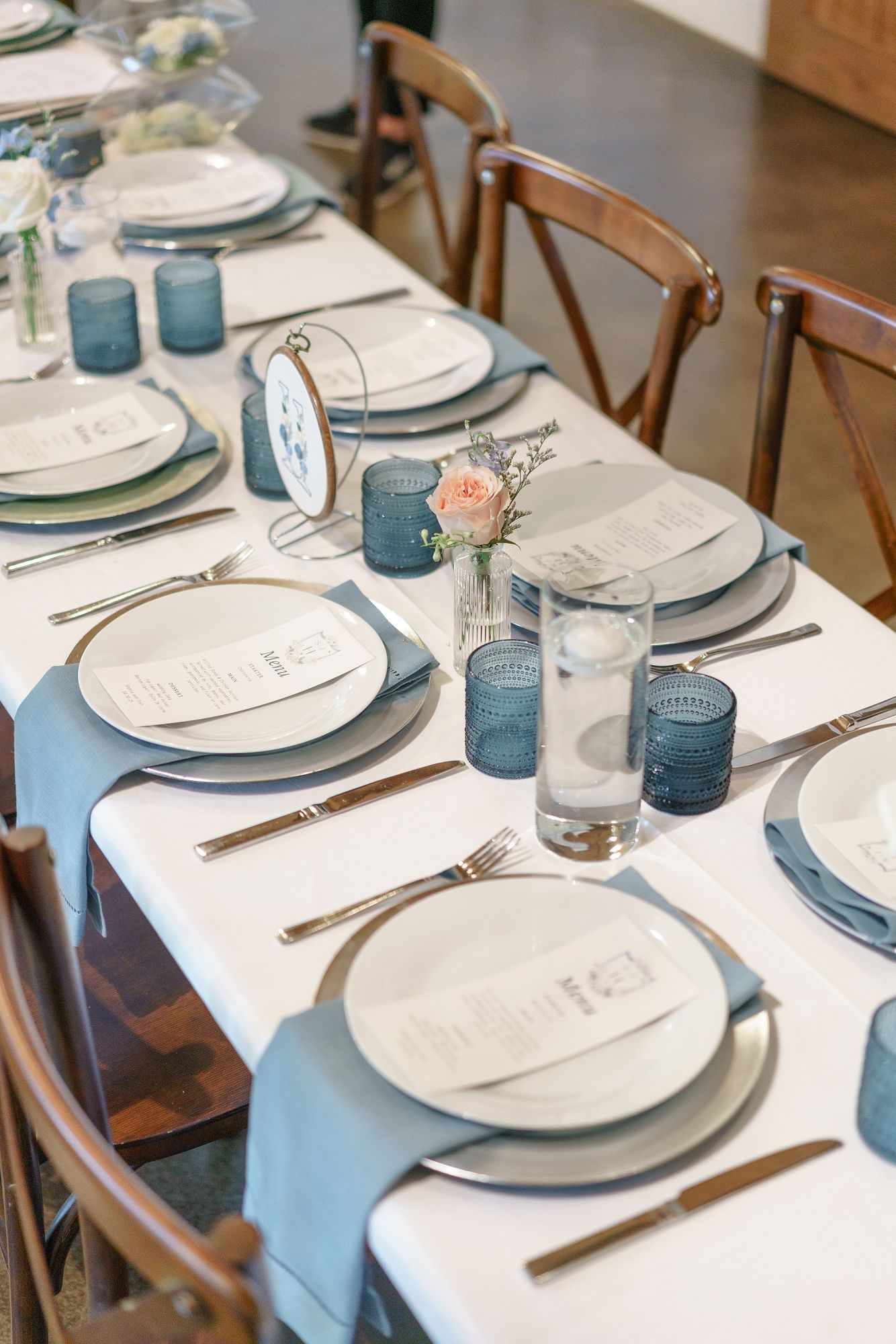 Elegant wedding table setting with blue glassware, white plates, silver chargers, and floral accents on a white linen tablecloth.