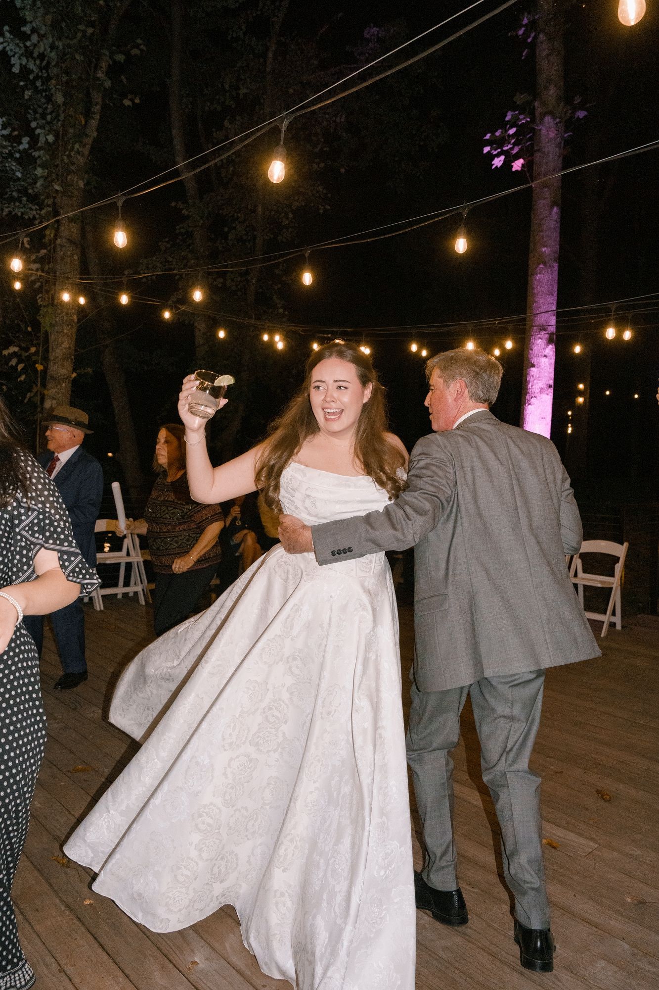 Bride in white ballgown dancing outdoors at night under string lights at wedding reception.