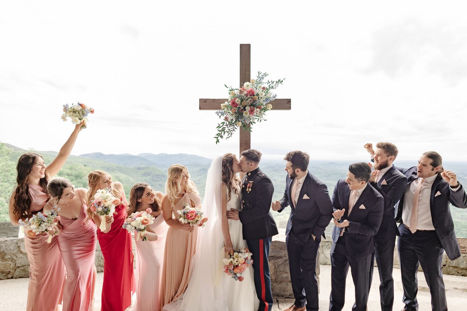 Wedding party celebrating before a wooden cross with mountain views, bridesmaids in pink and red gowns.
