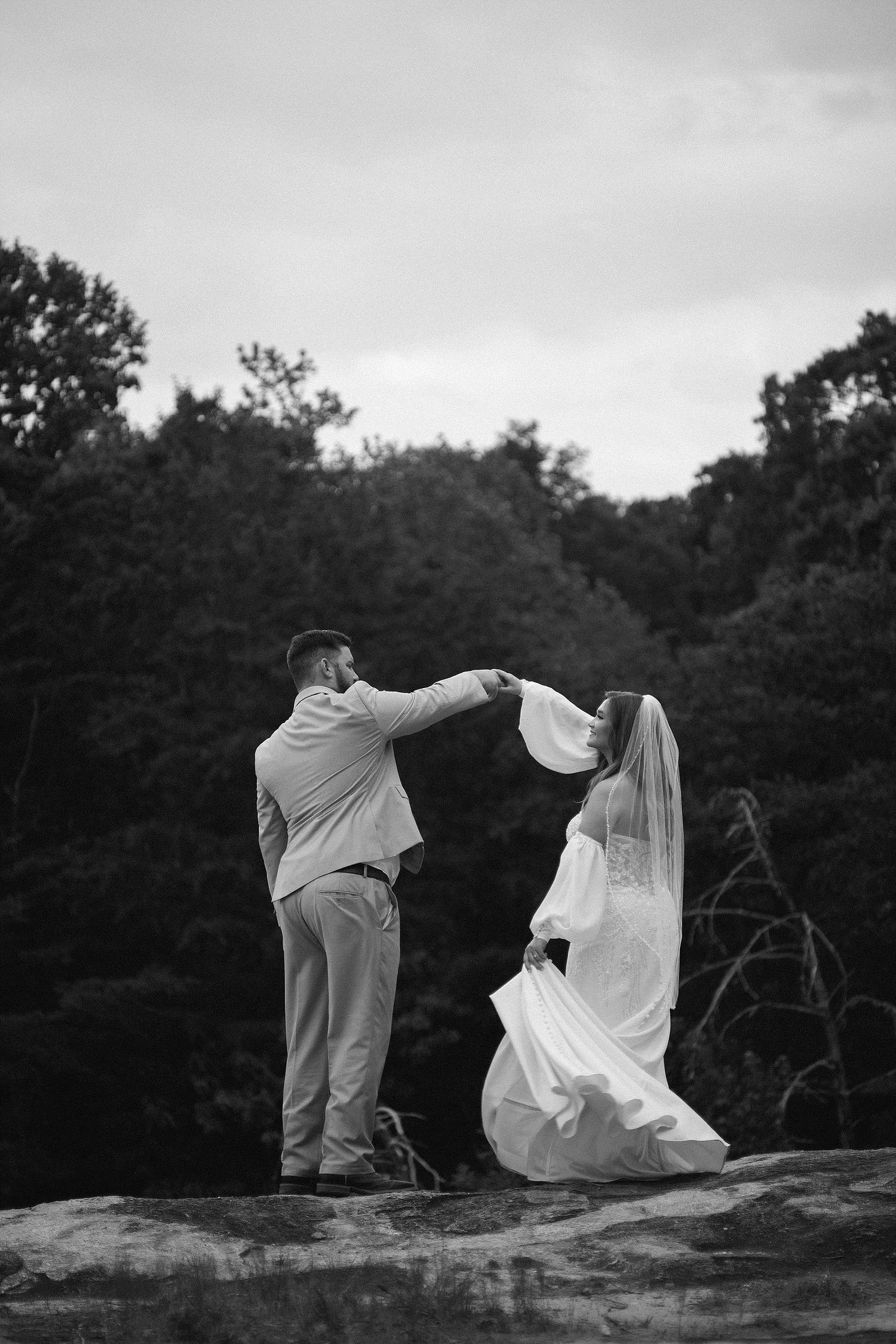 A black and white photo of newlyweds dancing on rocky mountain ledge.