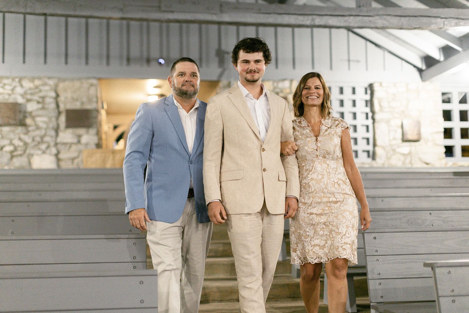 Three people in formal attire stand together on a porch with stone and wood features behind them.