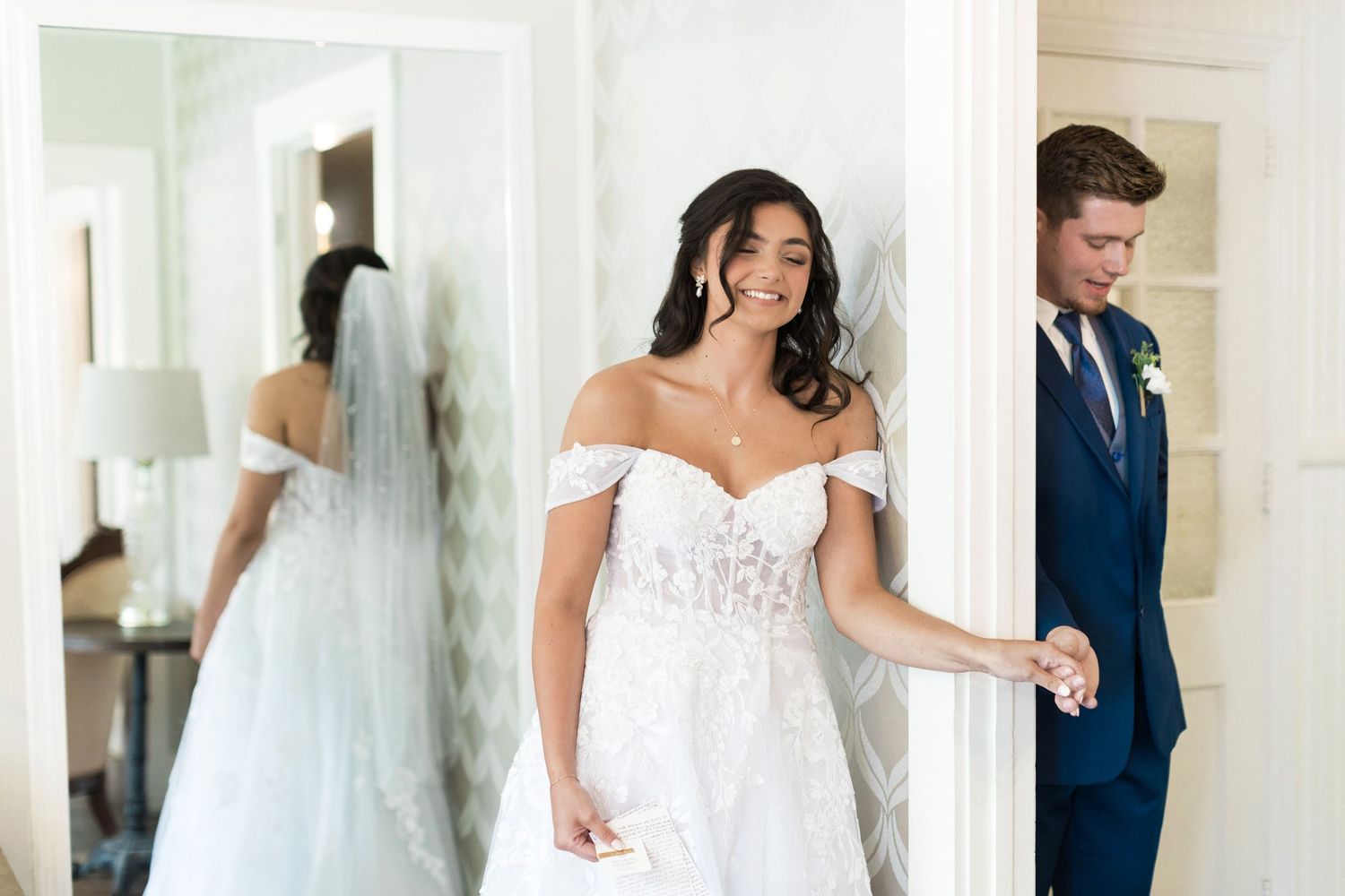 A bride in a white off-shoulder gown peeks around a corner while a groom in navy suit stands on the other side.