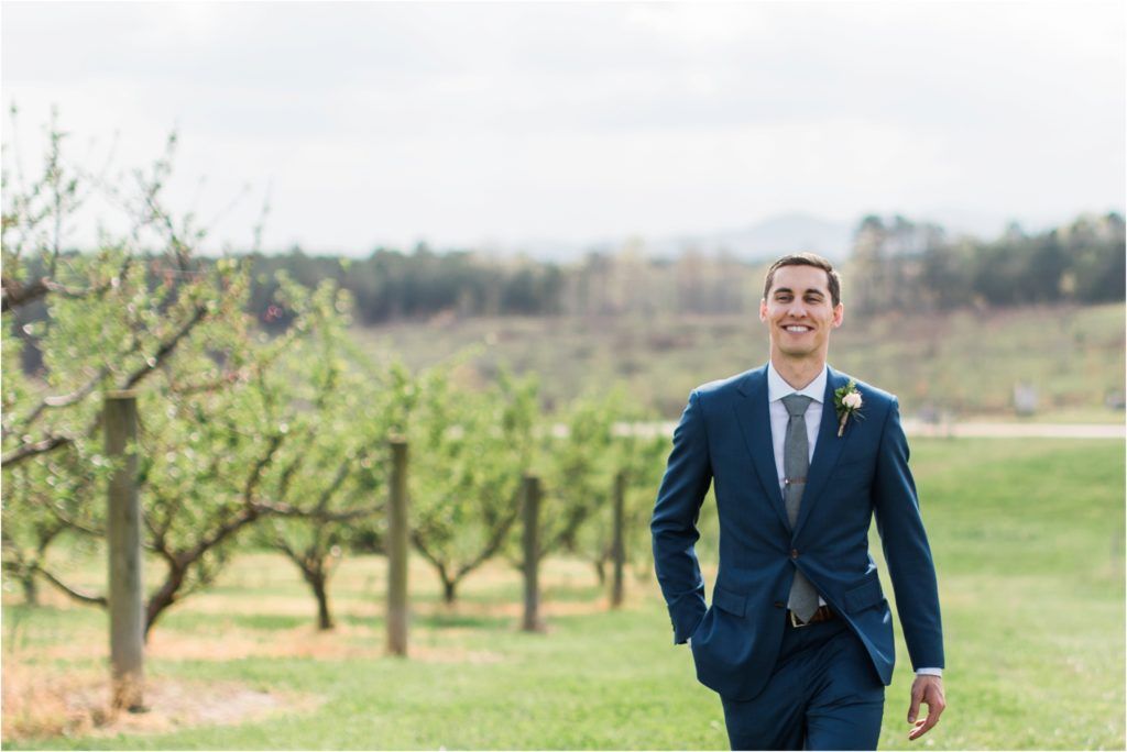 Groom in tailored navy suit standing in an orchard with rolling hills in background.