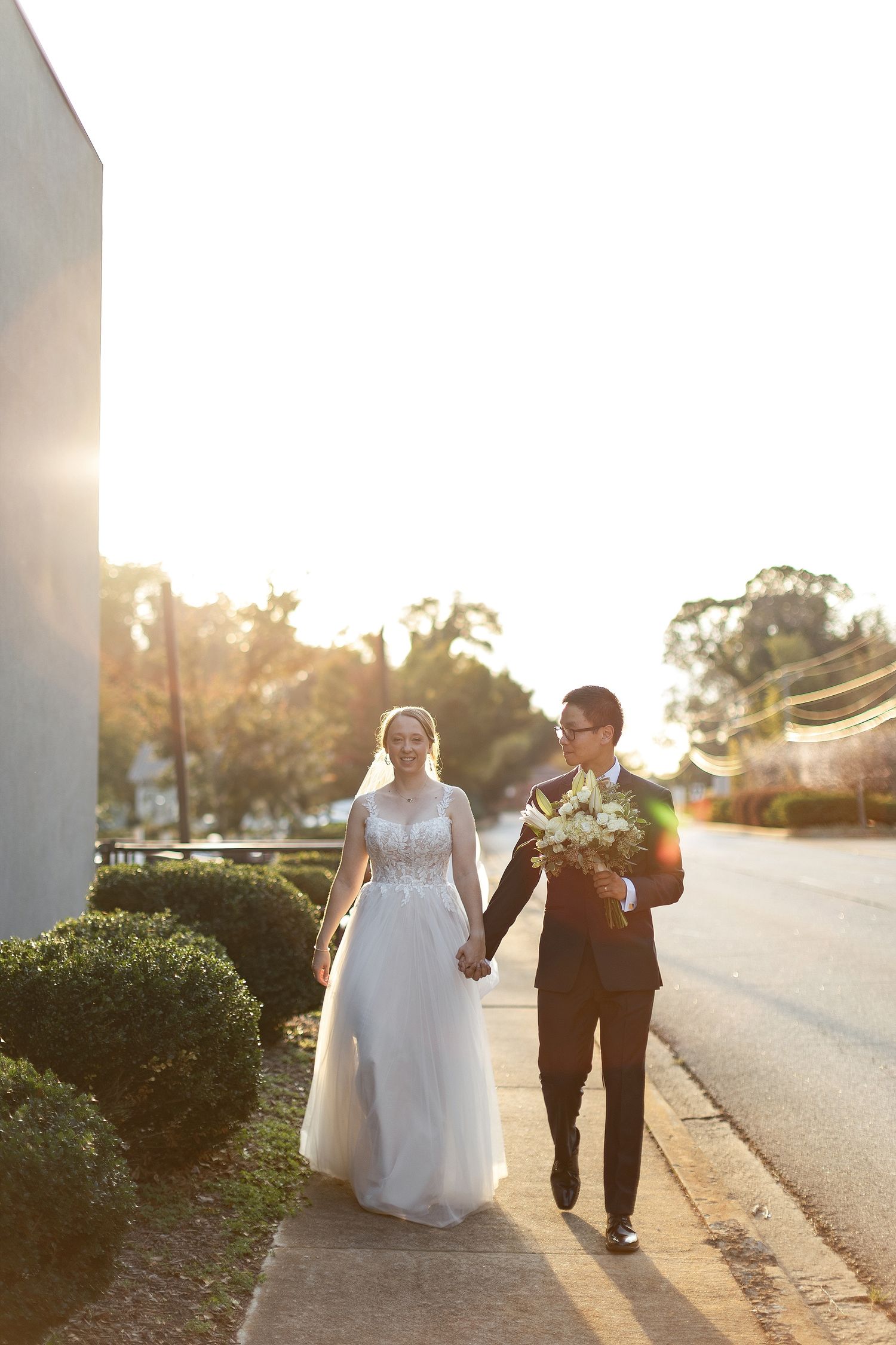 A couple walking hand in hand at sunset, with the bride wearing a white gown and groom in dark suit carrying flowers.
