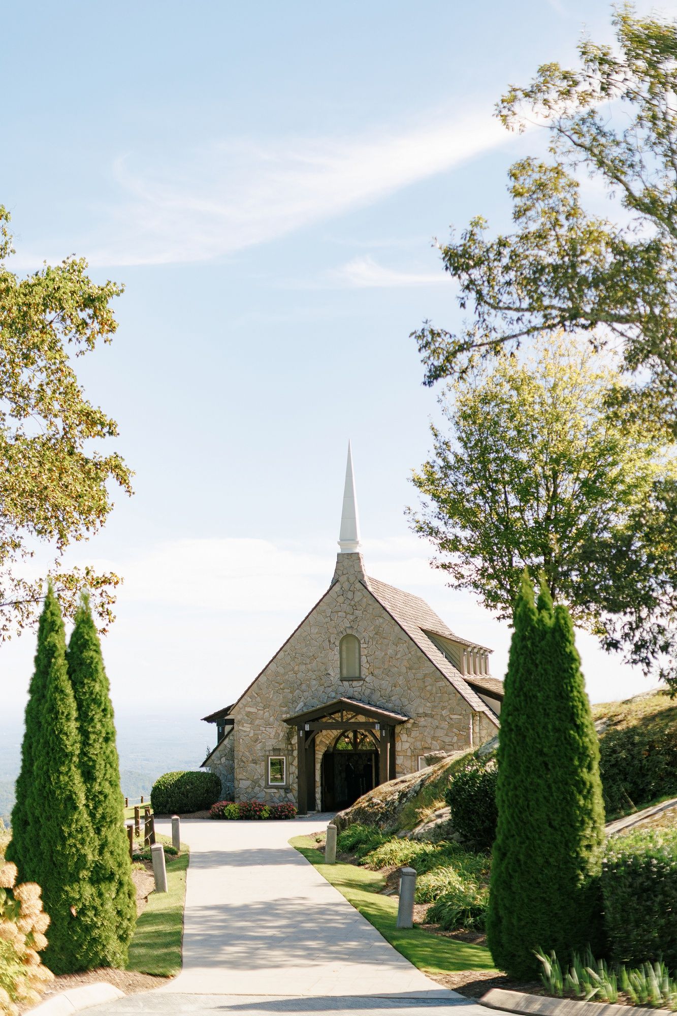 Stone church with steeple sits atop a hill surrounded by manicured evergreen trees and a winding driveway.