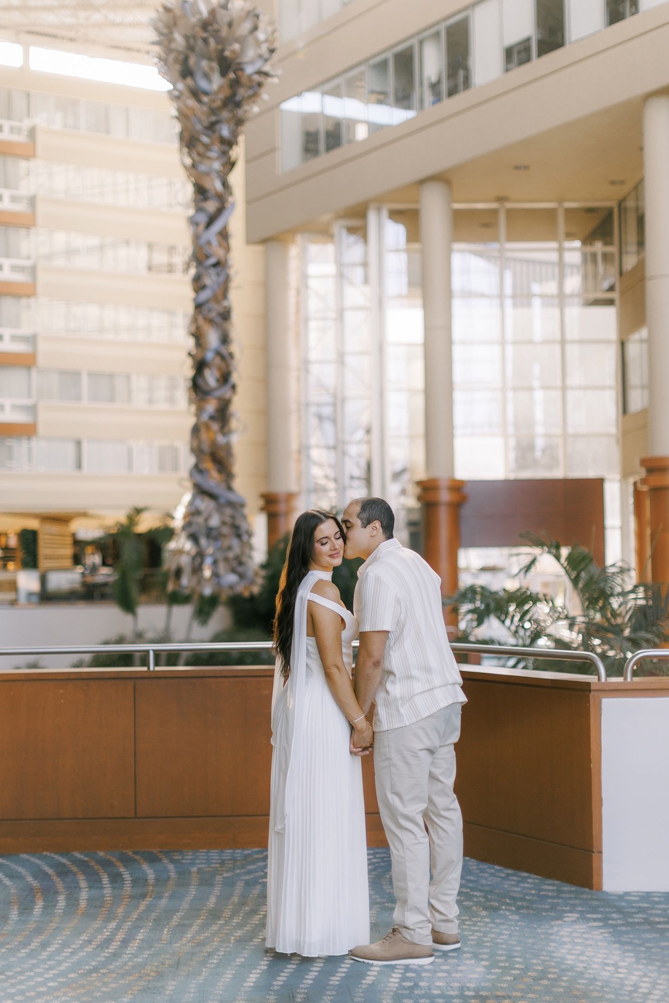 A couple shares a romantic moment in an elegant hotel lobby with palm trees and modern architecture.