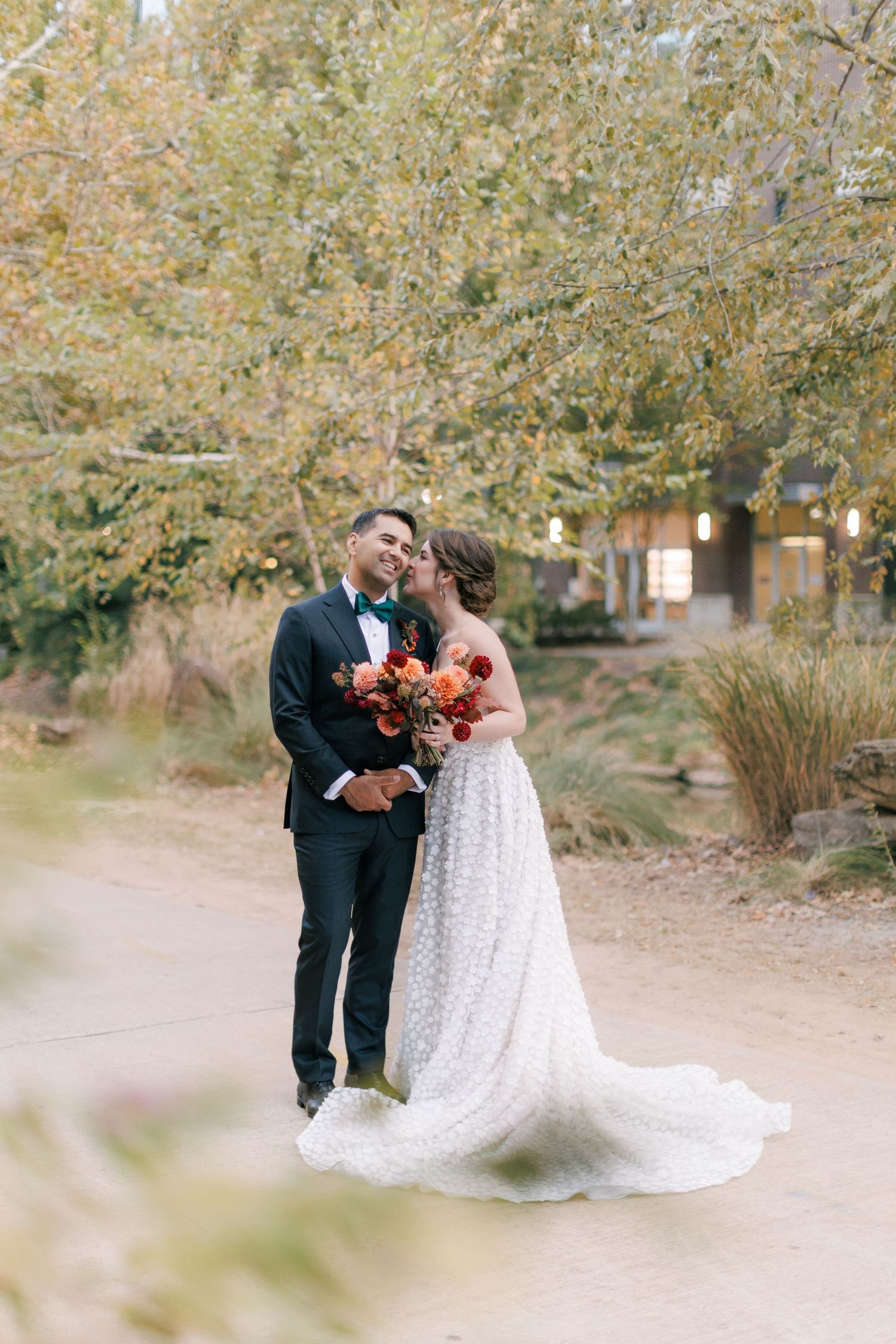 Bride and groom share a romantic moment outdoors, she holds a red bouquet, he wears a tuxedo, surrounded by autumn trees.