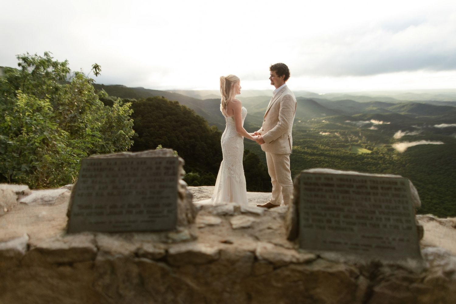 A couple holds hands during their mountaintop wedding ceremony overlooking a vast valley landscape at sunset.
