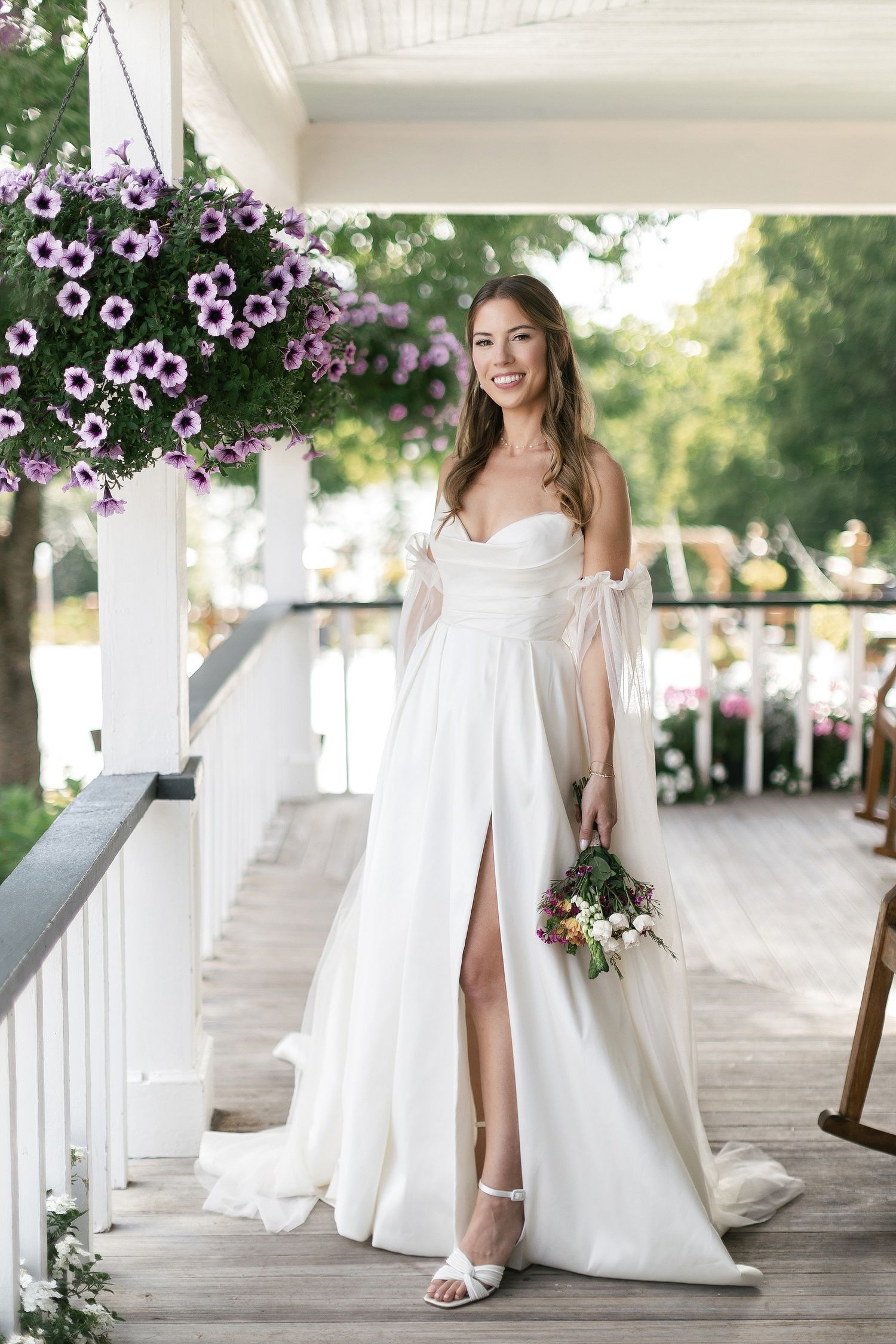 A bride in an elegant white gown with slit poses on a porch decorated with purple flowers.